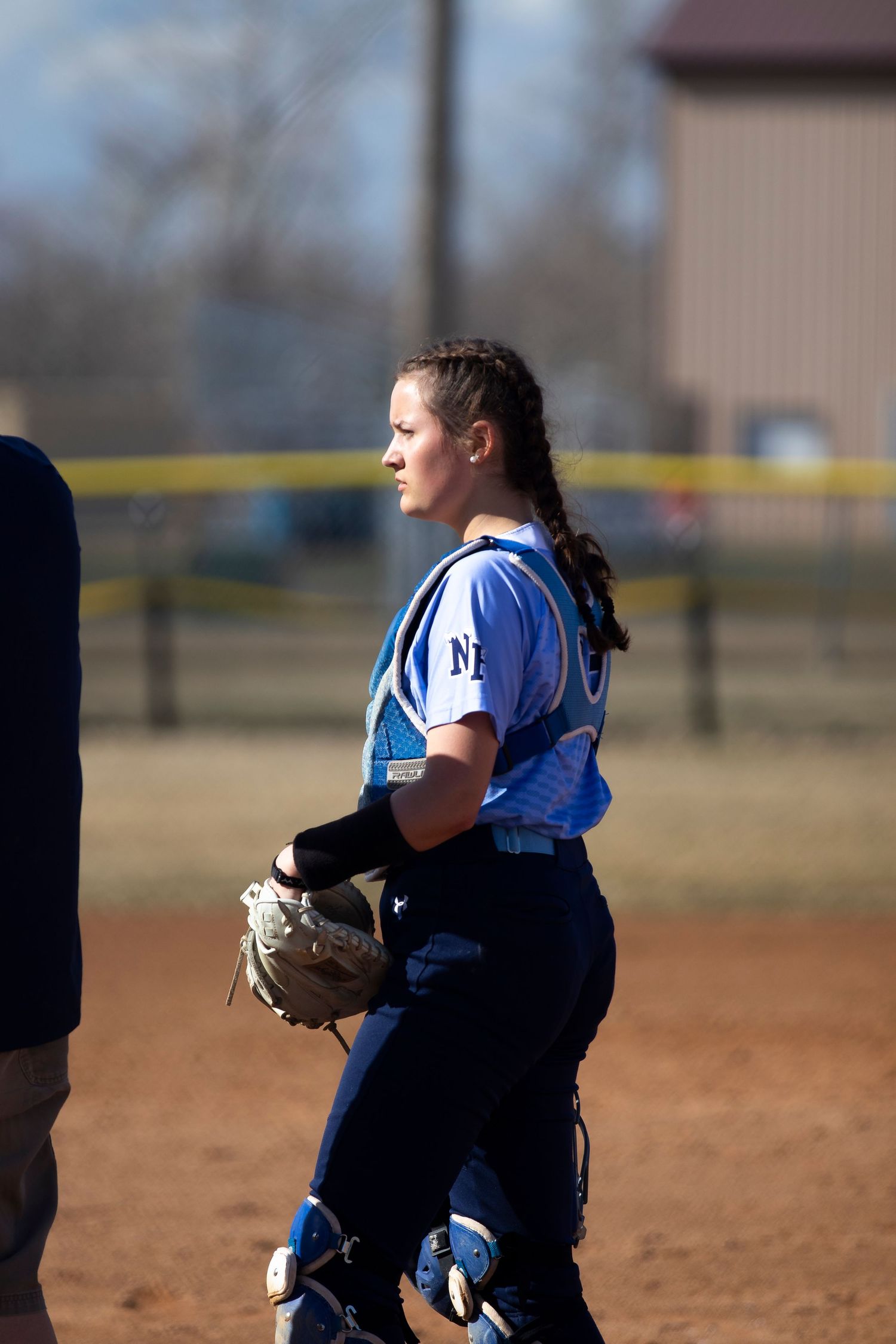Female softball catcher in blue uniform holding glove on dirt field during a game.