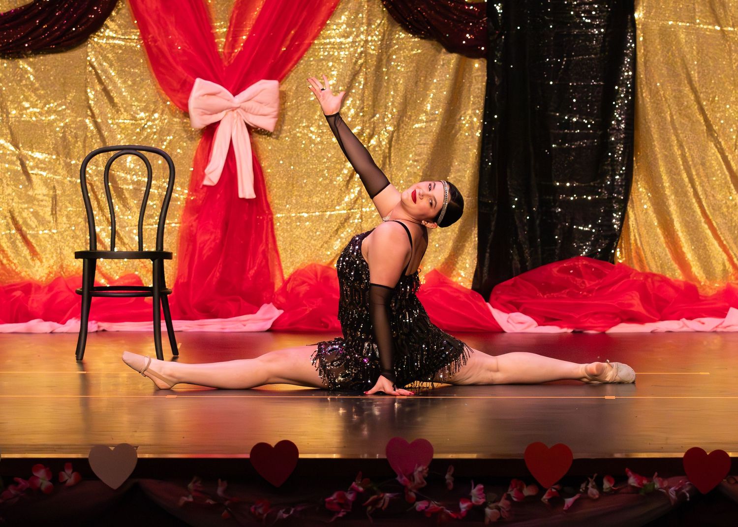 Dancer in black sequin costume performs a full split on stage floor with gold sequin backdrop and chair.