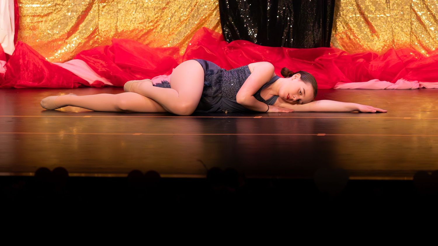 A dancer lies gracefully on a wooden stage floor in a blue costume, surrounded by vibrant red and gold fabric backdrops.