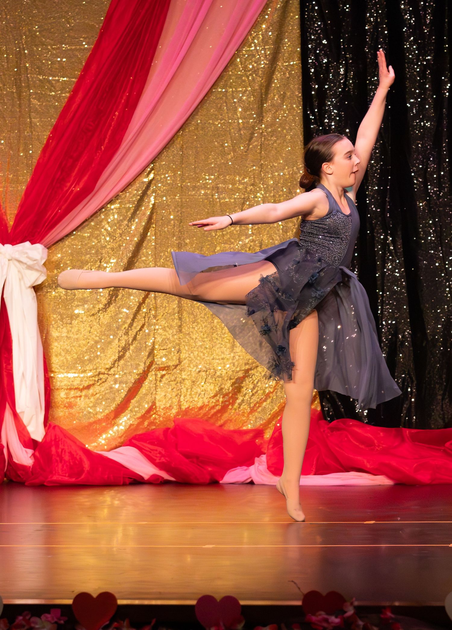 A graceful dancer leaps on stage in a gray tulle dress against a gold sequin and red drape backdrop.