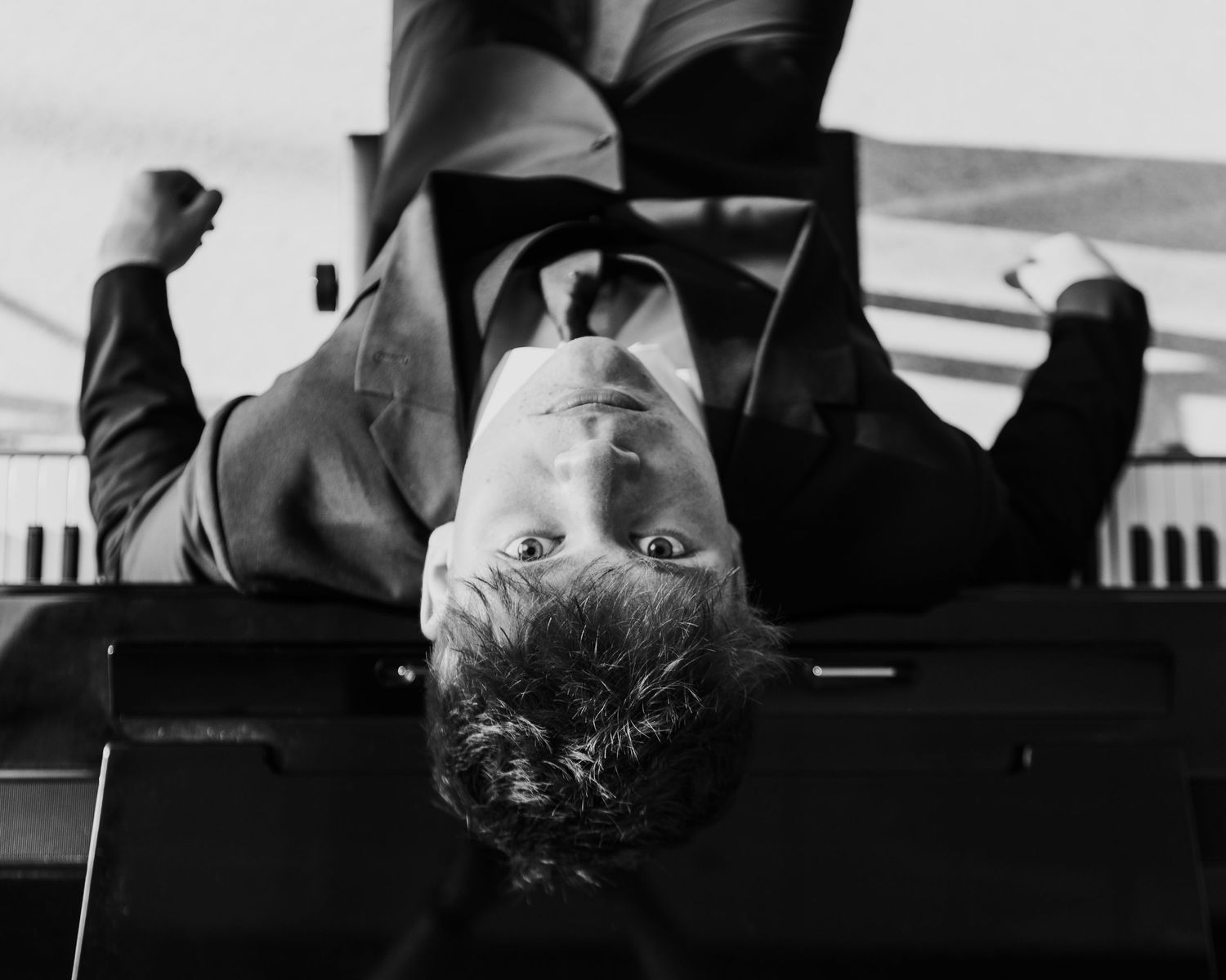 Black and white photo of a man in a suit lying upside down near a piano, looking up with wide eyes.
