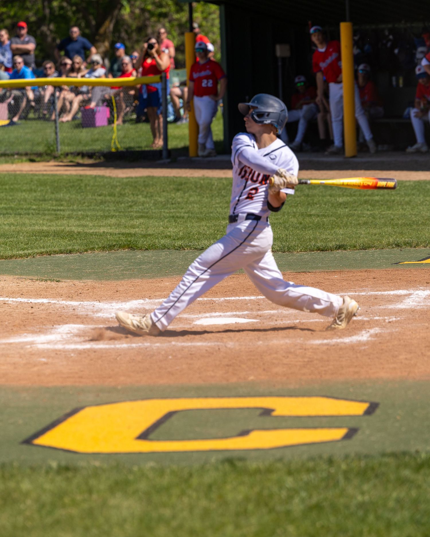 Young baseball player in white uniform swinging a yellow bat during a sunny youth league game with spectators watching.