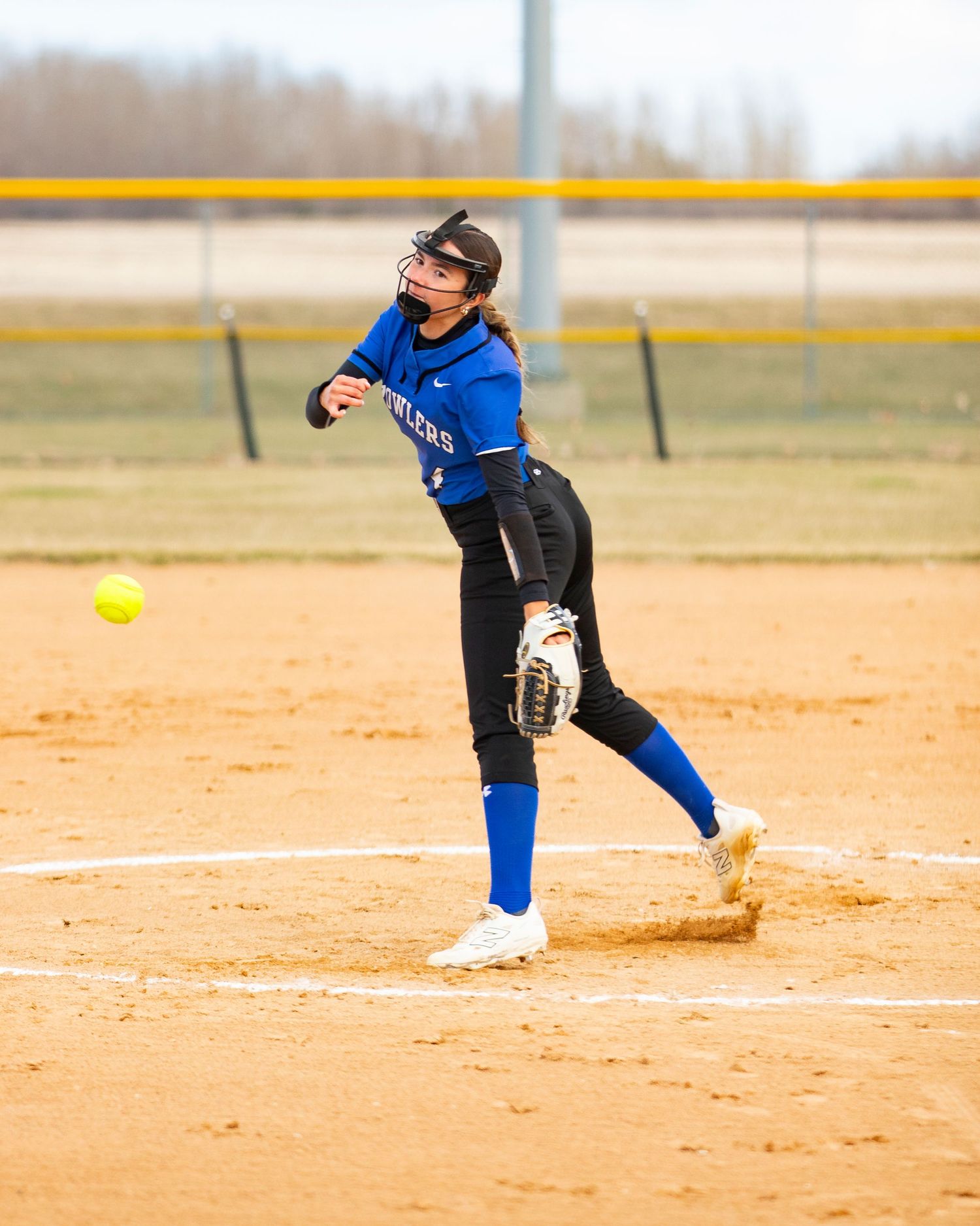 Softball pitcher in blue uniform mid-windup releasing yellow ball on dirt mound during game.