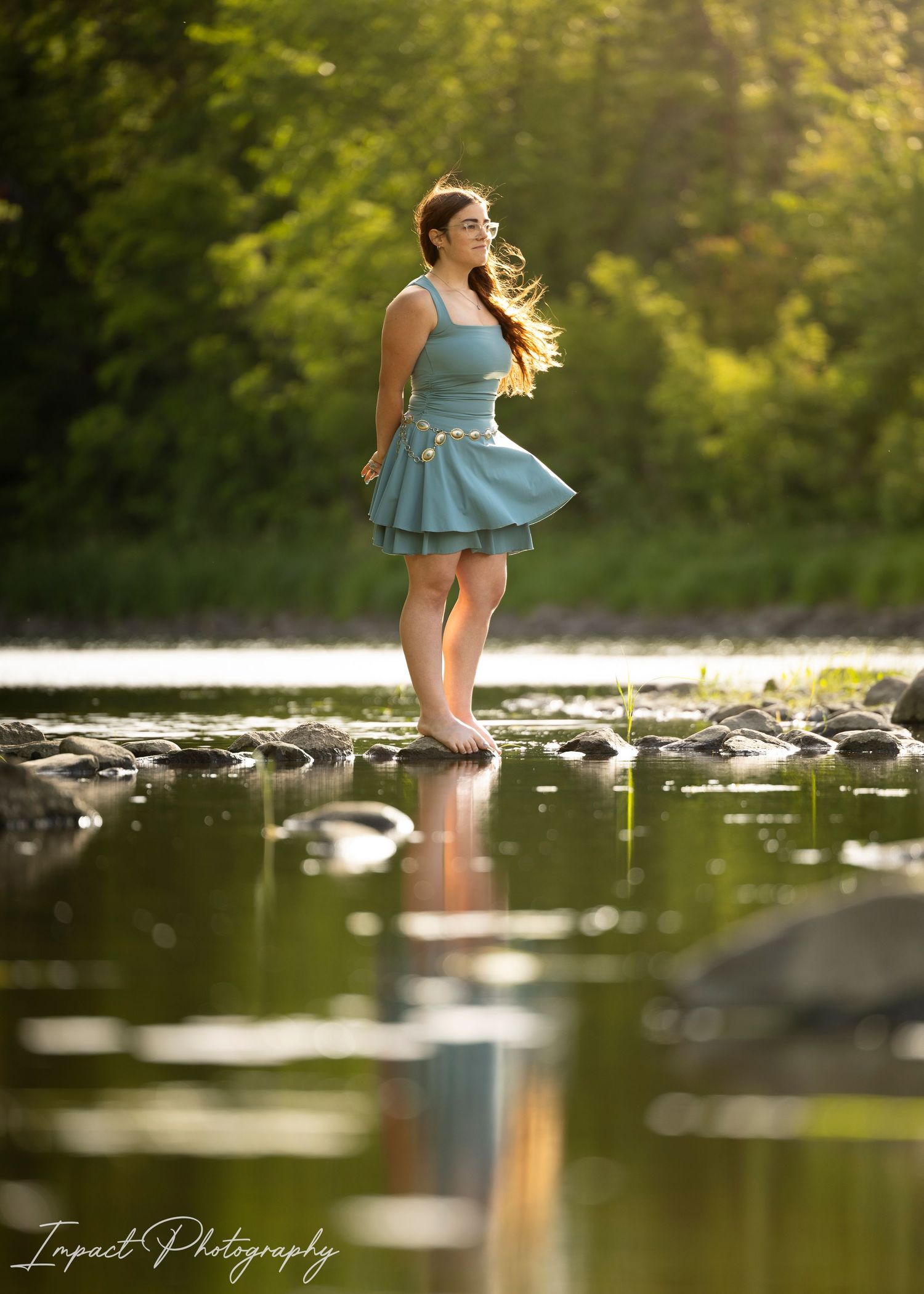 Young woman in teal dress standing on river rocks, hair flowing, with lush green trees and golden light reflection.