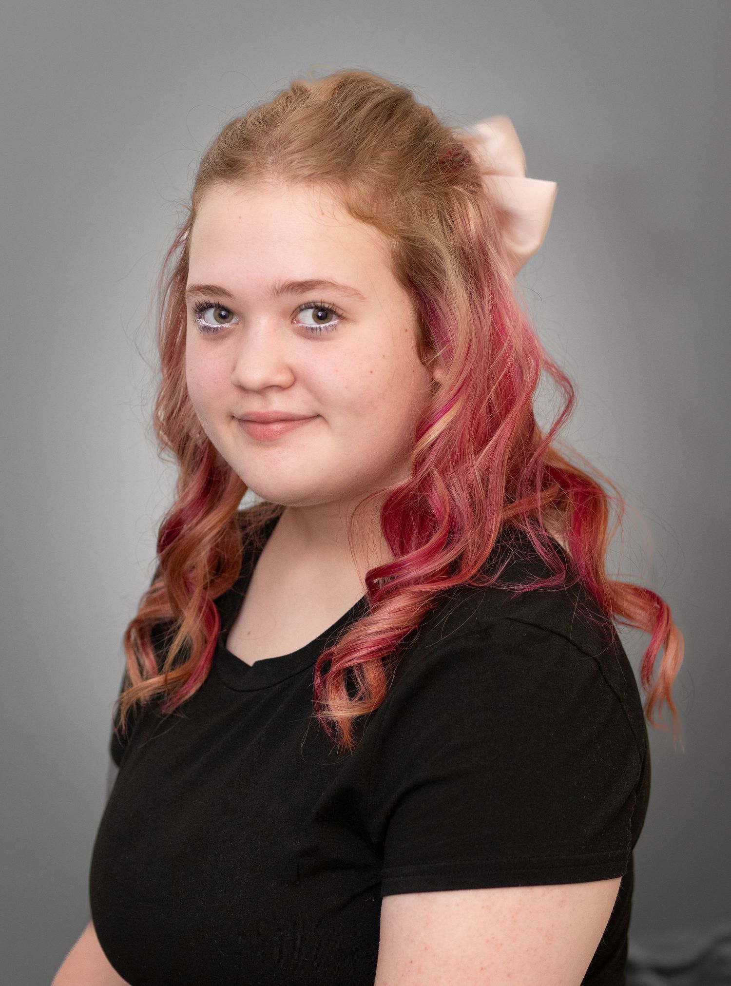 Young woman with pink and blonde curly hair and a bow accessory wearing a black t-shirt.