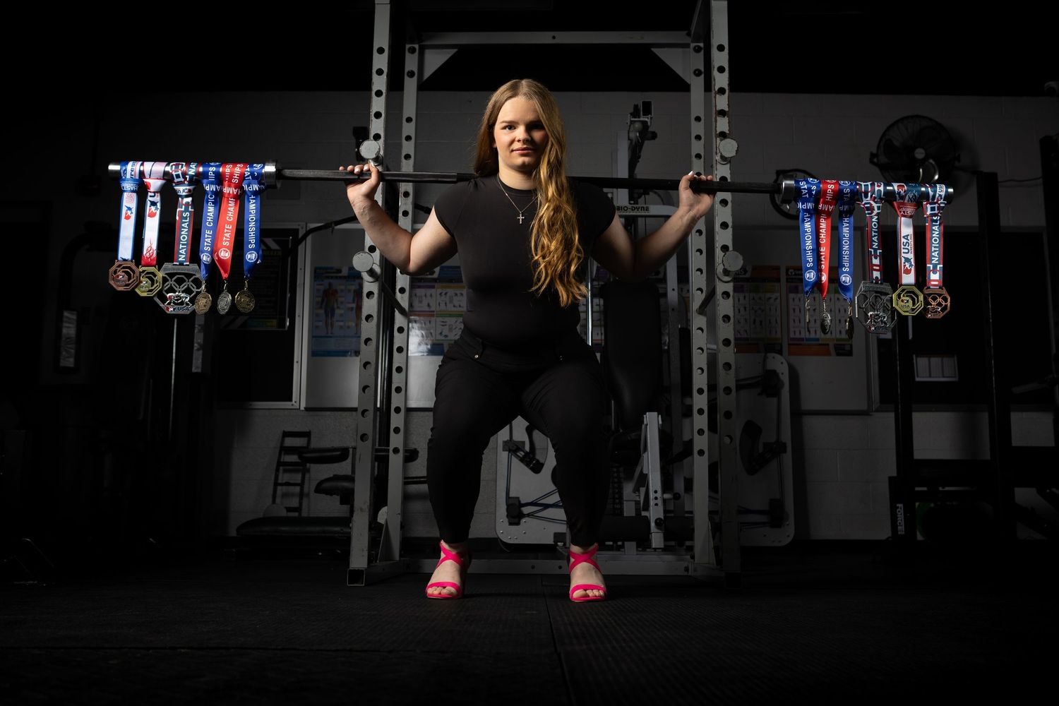 Female powerlifter squatting with barbell in dark gym surrounded by colorful competition medals on display rack.