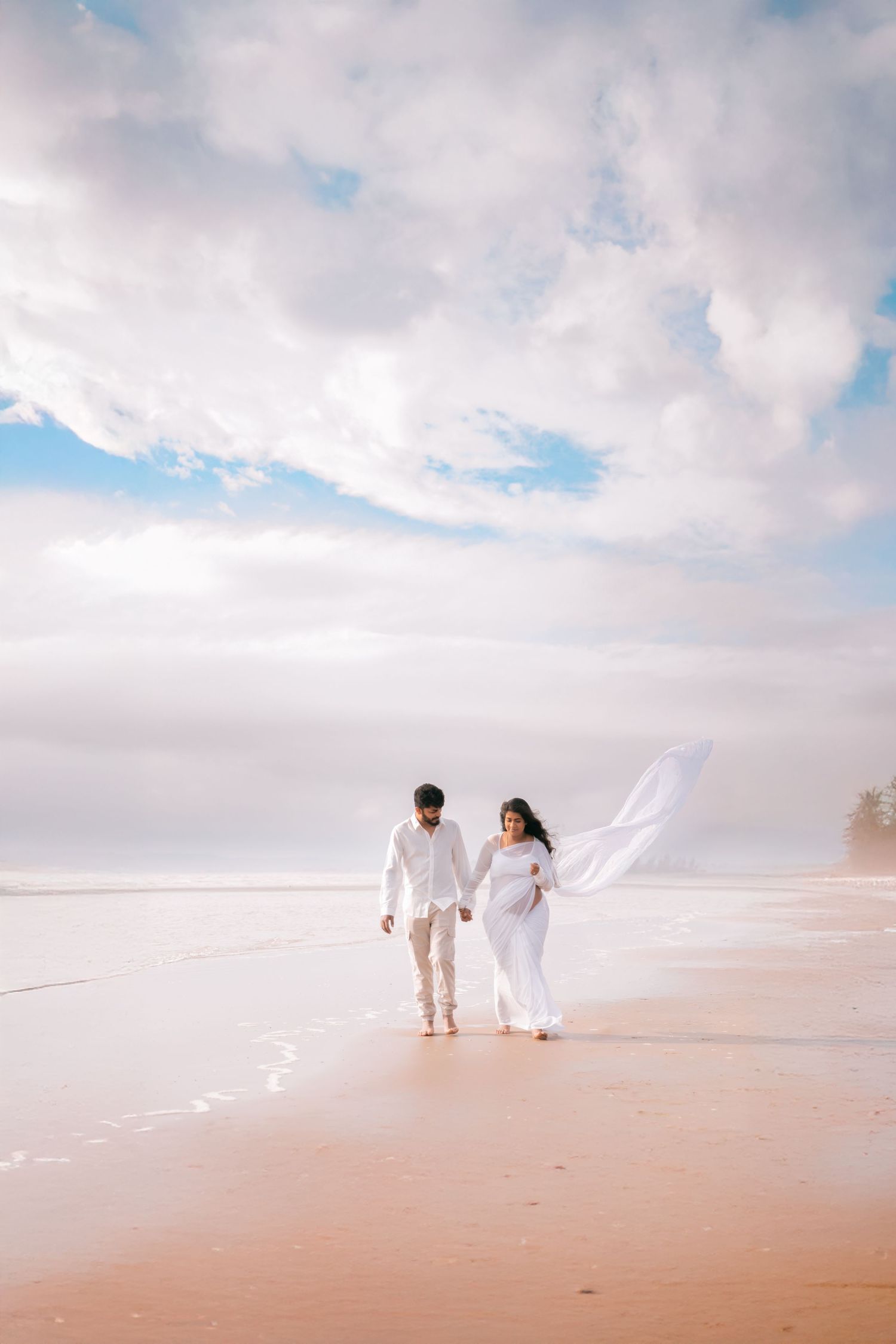 Pregnant couple in white outfits walking hand-in-hand on a sandy beach with dramatic cloudy sky.