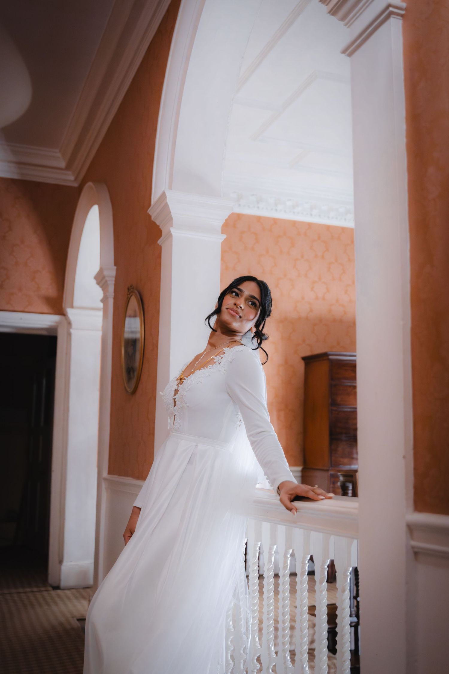 Elegant bride in long-sleeve white wedding gown standing gracefully on a balcony in a classic interior.