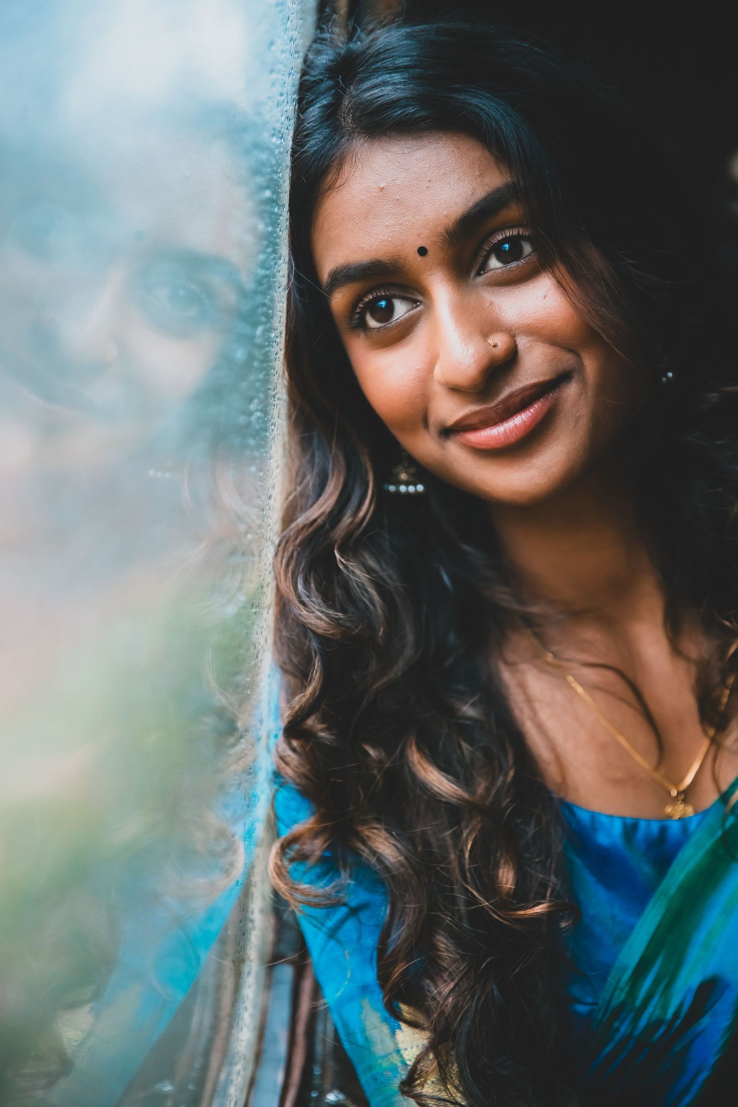 A smiling young woman in a blue saree with a bindi leans against a window frame.