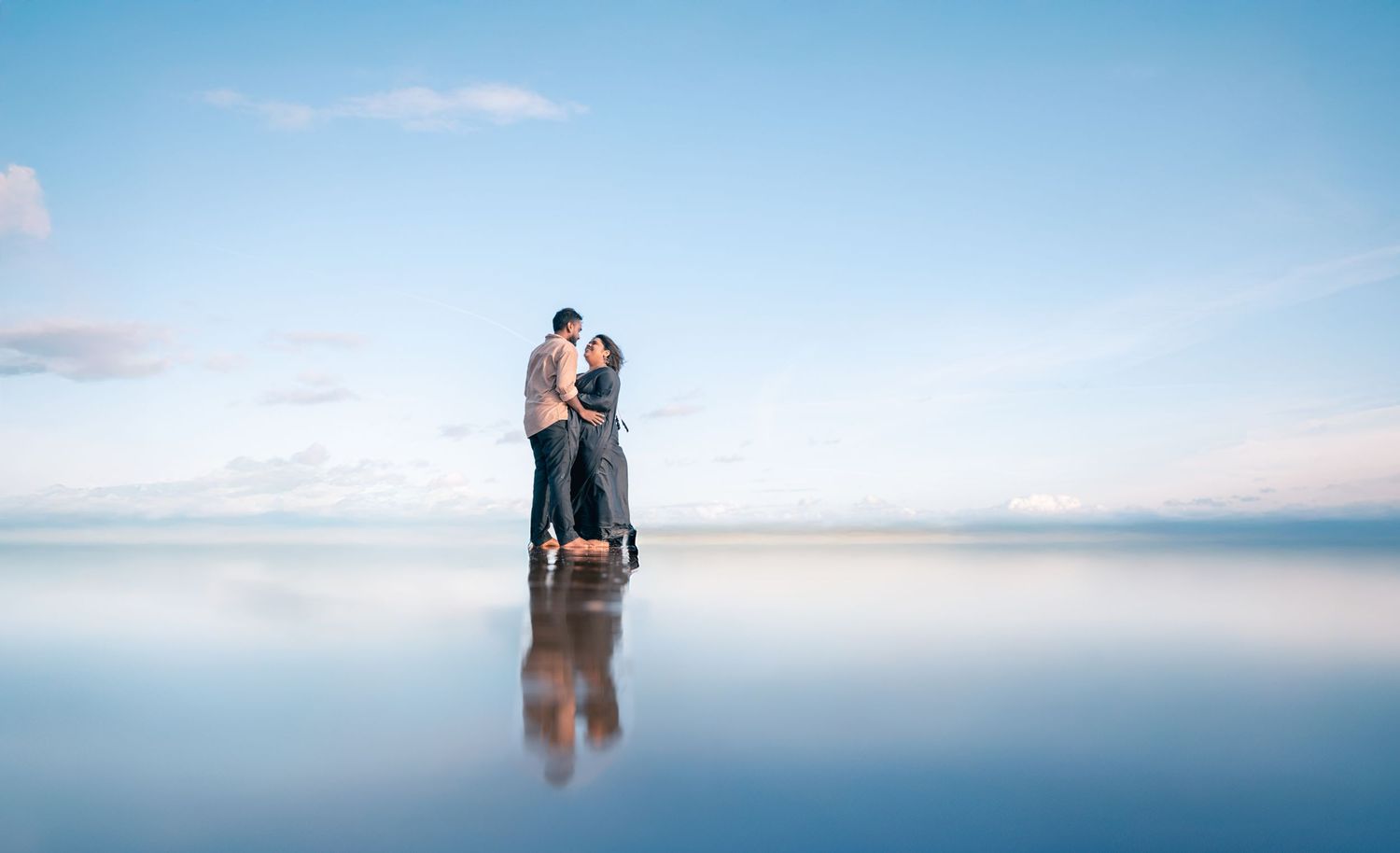 Couple hugging on a reflective wet surface under a clear blue sky, creating a mirror effect.