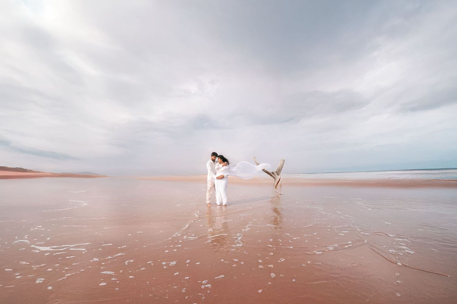 Bride in white dress twirling veil on a serene pink sandy beach.