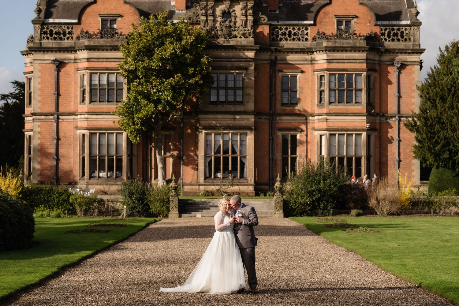 Bride and groom embrace on gravel path before grand red brick Victorian manor house wedding venue.