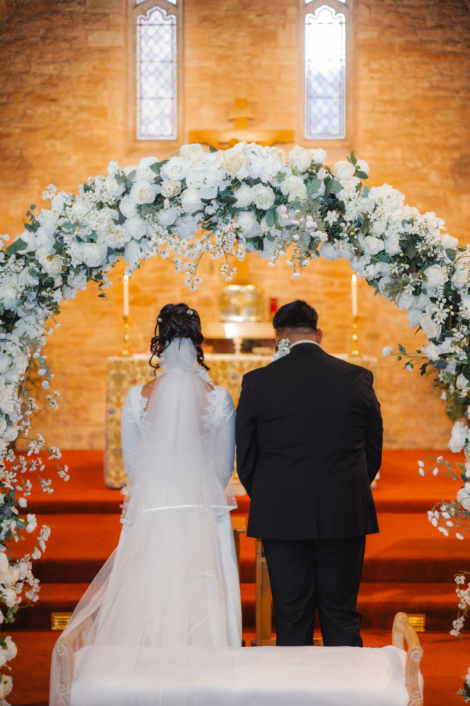 Bride and groom standing at church altar beneath white floral arch during wedding ceremony.