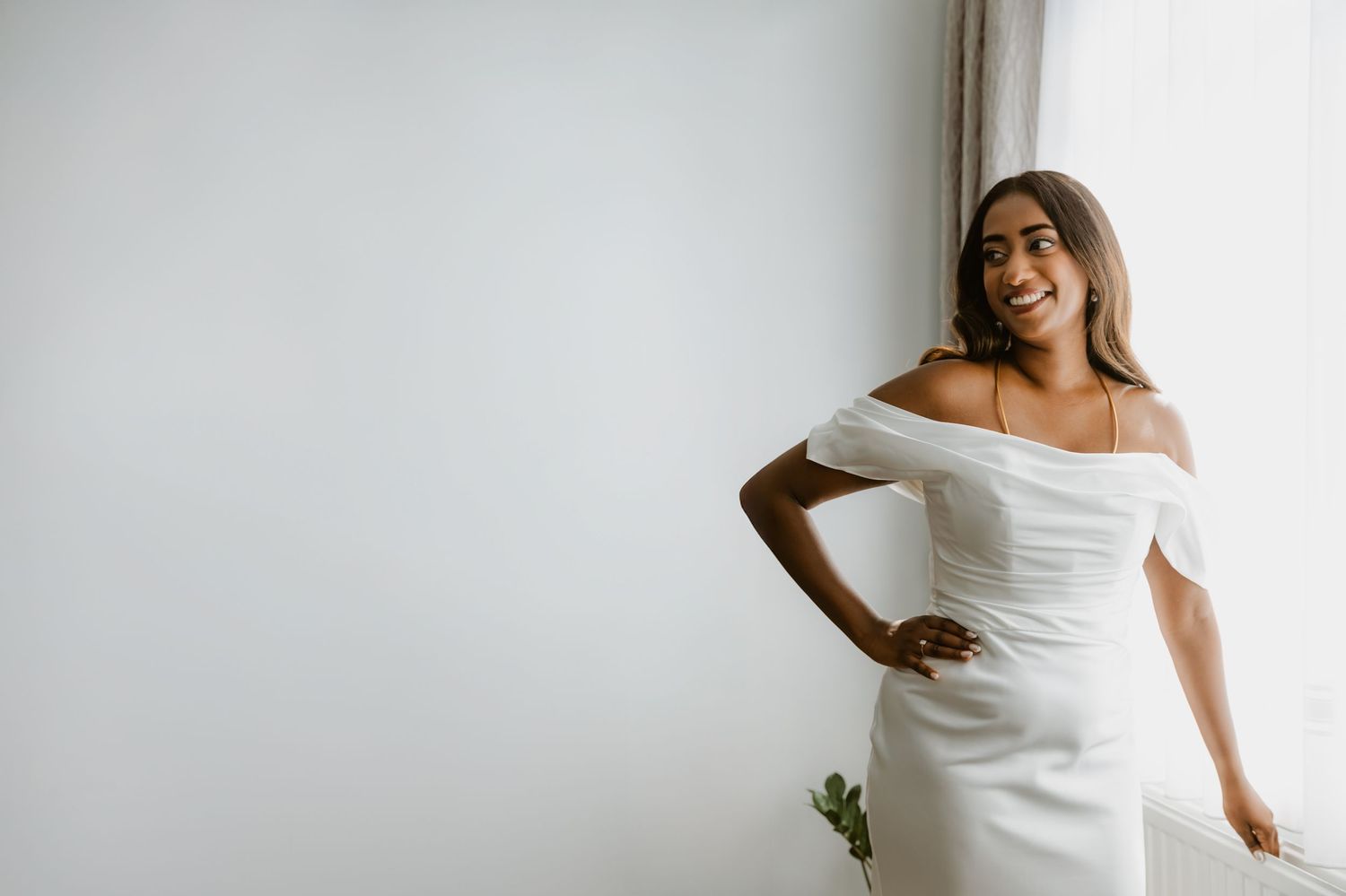 Smiling bride in off-shoulder white wedding dress posing by a bright window.