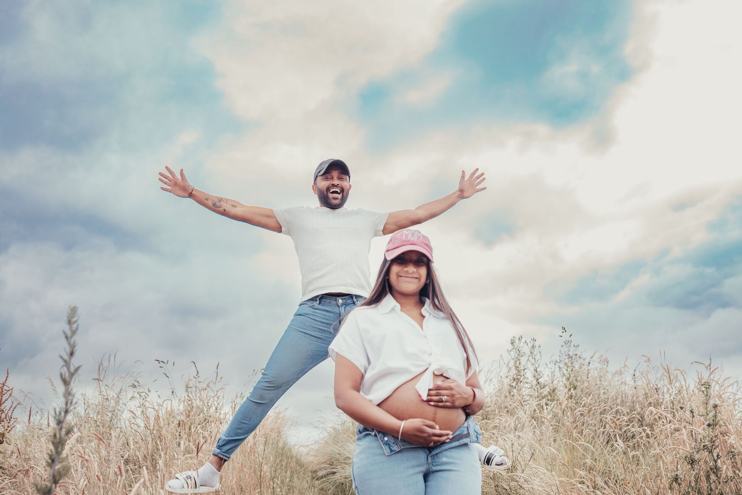 Playful maternity photo of couple in white outfits in a field, man posing with arms outstretched.
