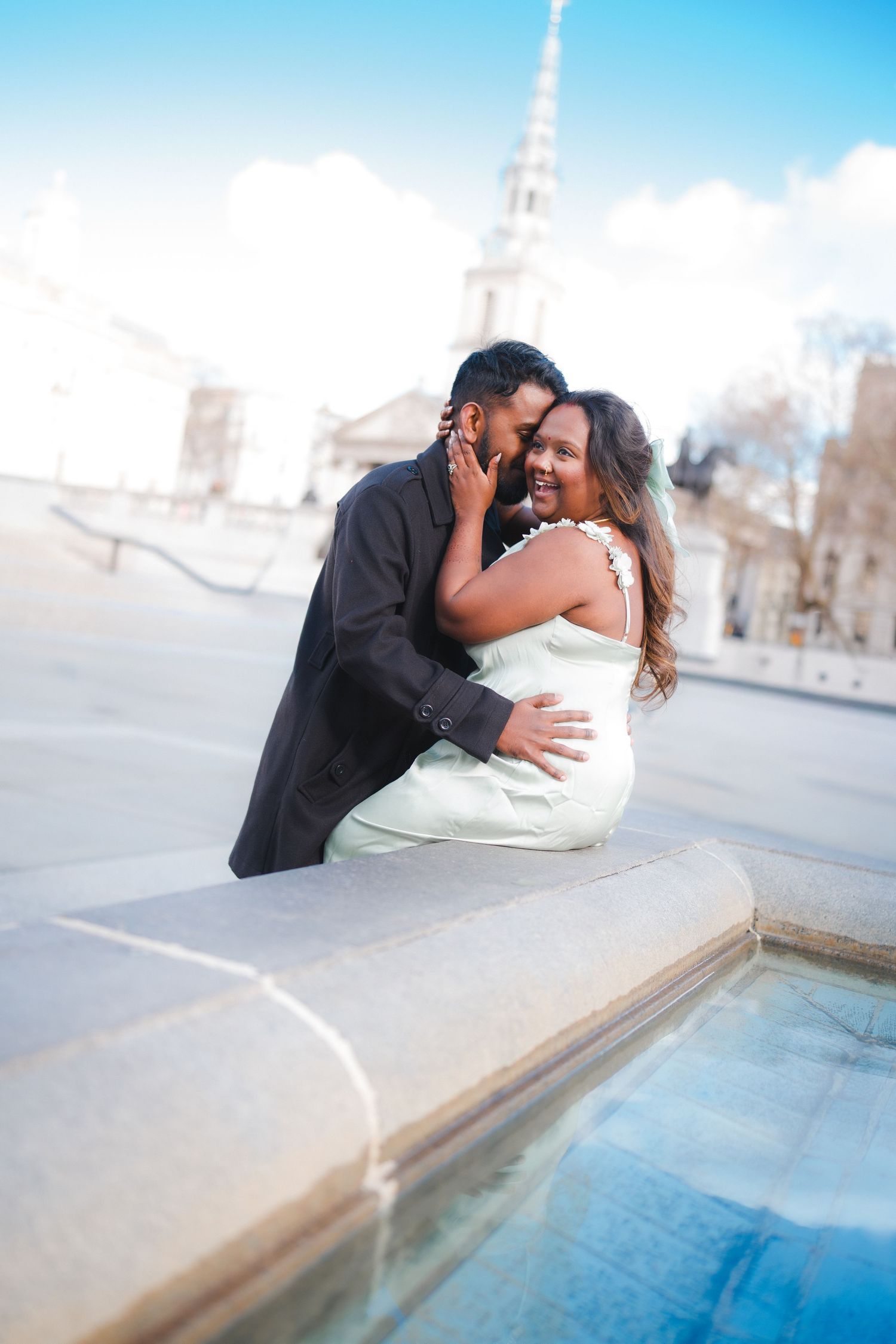 Couple sharing an intimate moment by a fountain with a church steeple in the background.