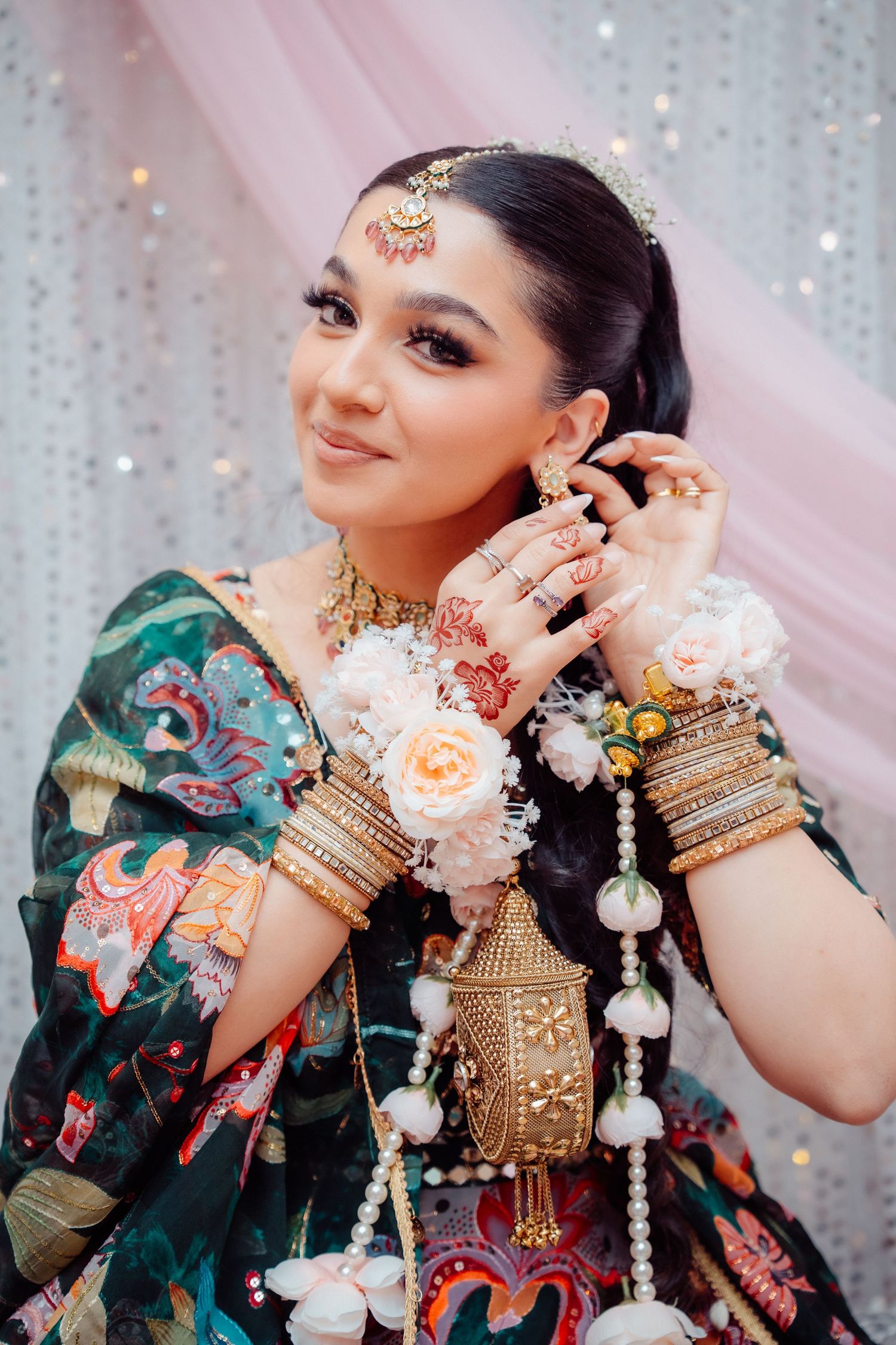 A bride in a green floral lehenga adorned with gold bangles, maang tikka, and floral wrist accessories poses gracefully.