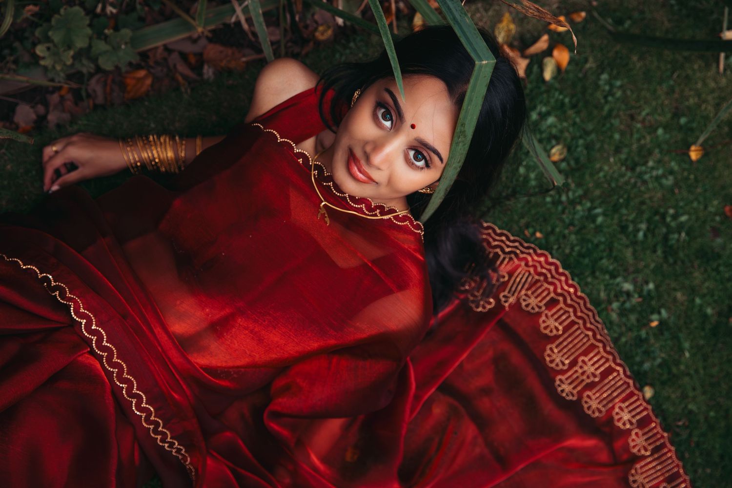 Beautiful Indian bride in red saree lying on grass with bindi and gold bangles in nature.