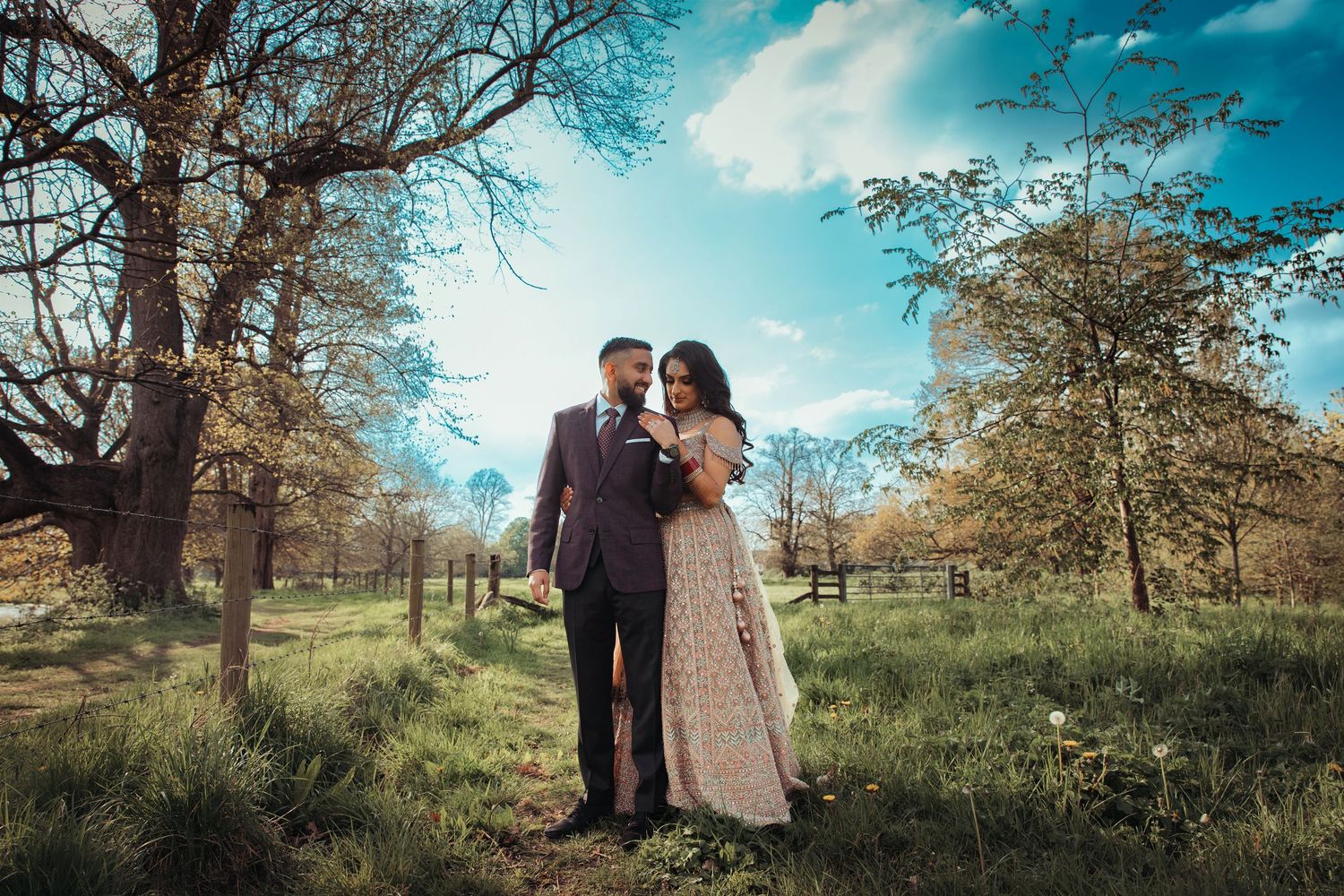 Couple embracing in a sunlit park with bare trees and green grass under a blue sky.