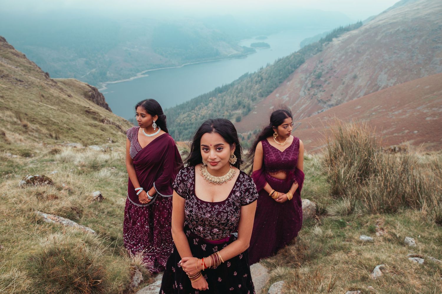 Three women in elegant purple Indian attire pose on a scenic mountain trail with a lake valley backdrop.