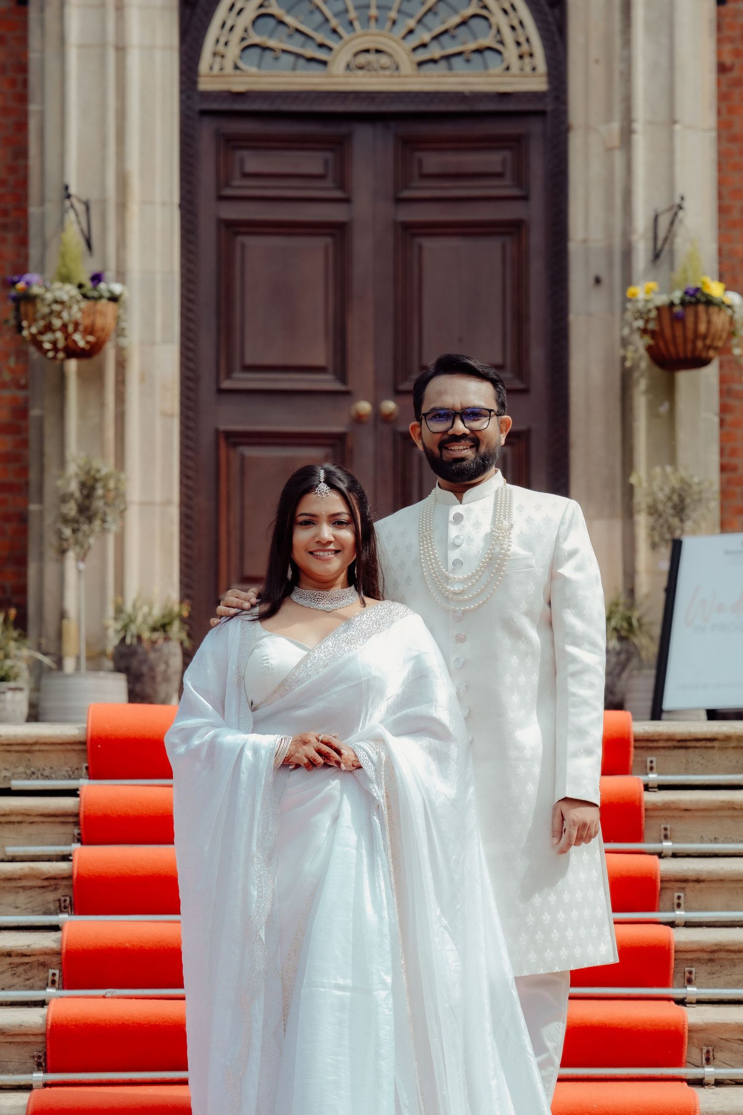 A couple in white traditional Indian attire poses on red-carpeted steps before a grand wooden door.