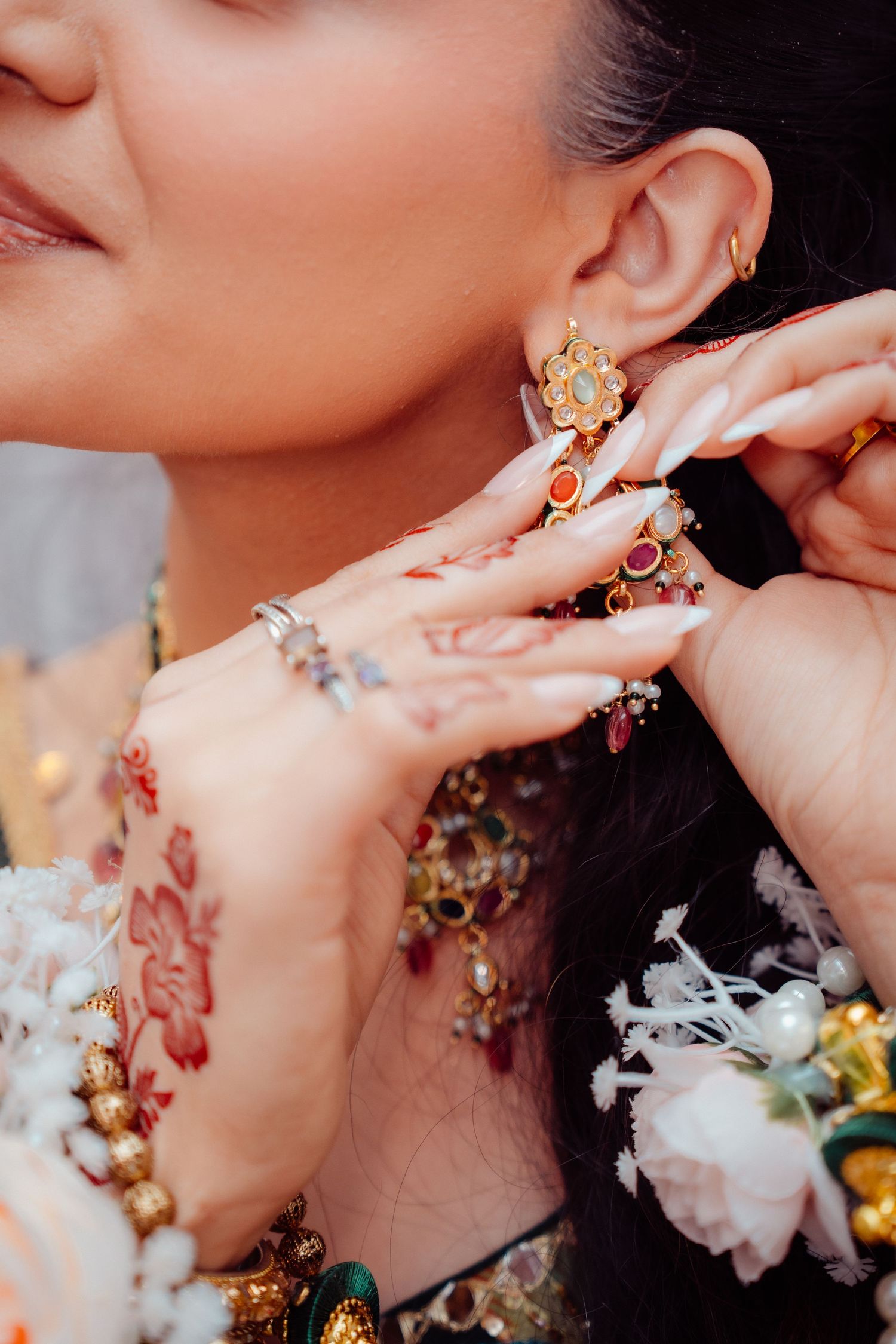 Close-up of a bride adjusting her gold floral earring, adorned with henna, long nails, and colorful jewelry.