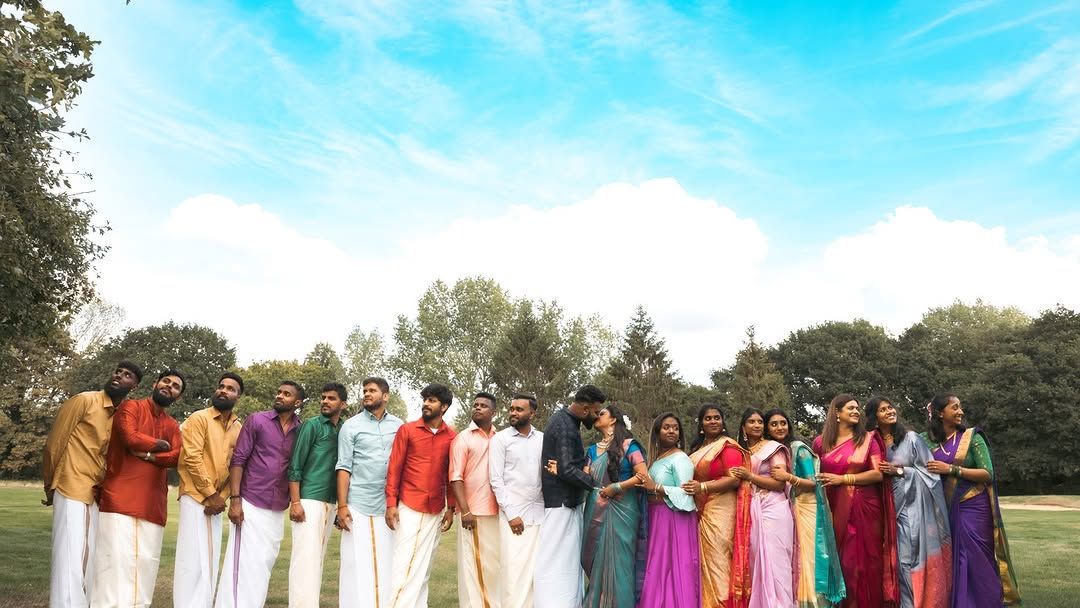 A large group of people in vibrant traditional Indian attire pose outdoors on green grass under a bright blue sky.