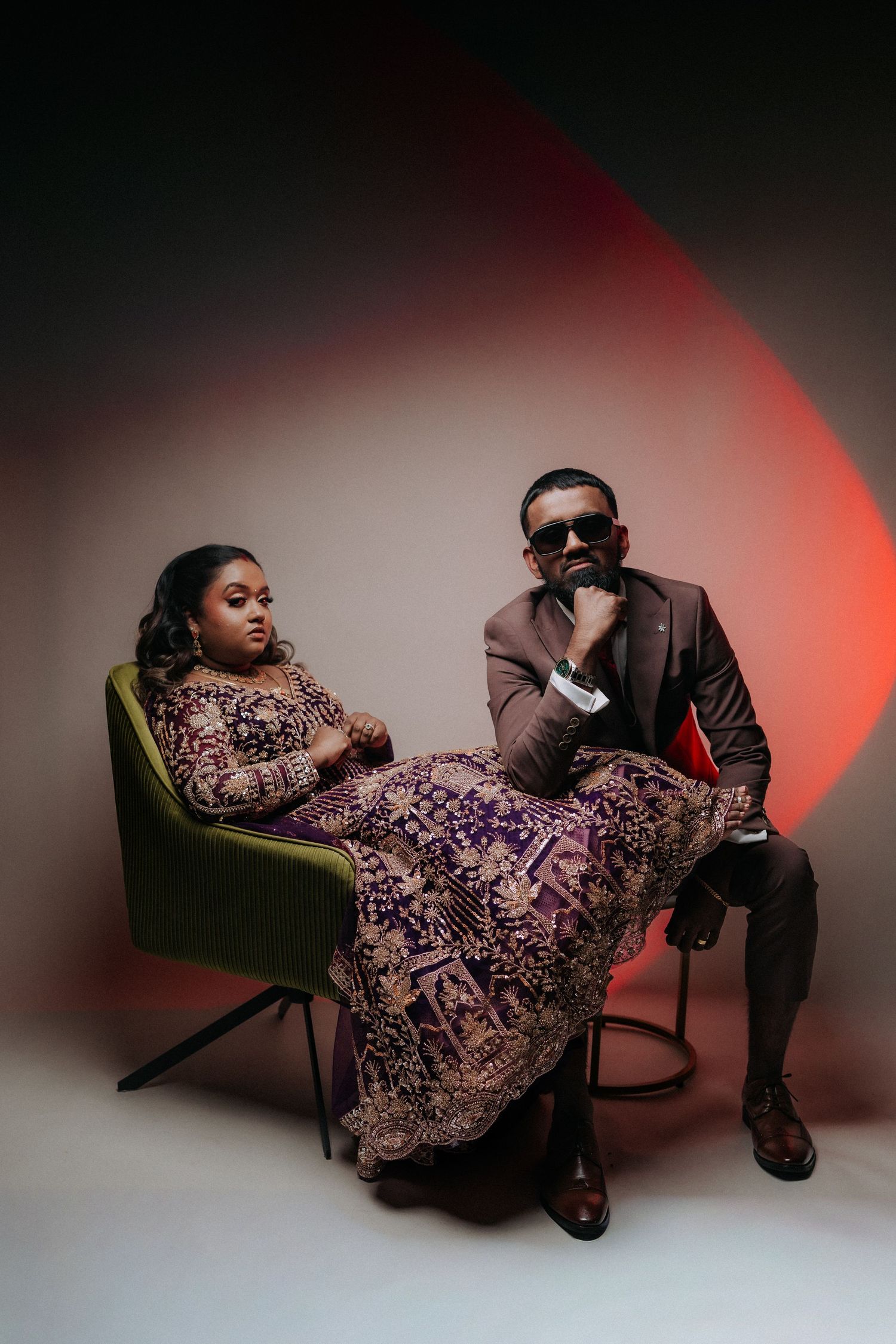 Elegant couple posing in studio, woman in purple embroidered lehenga, man in brown suit.