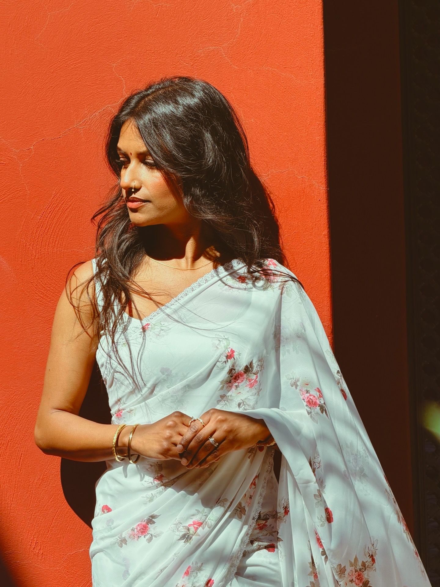 Indian woman in light floral saree standing against a vibrant red wall in sunlight.