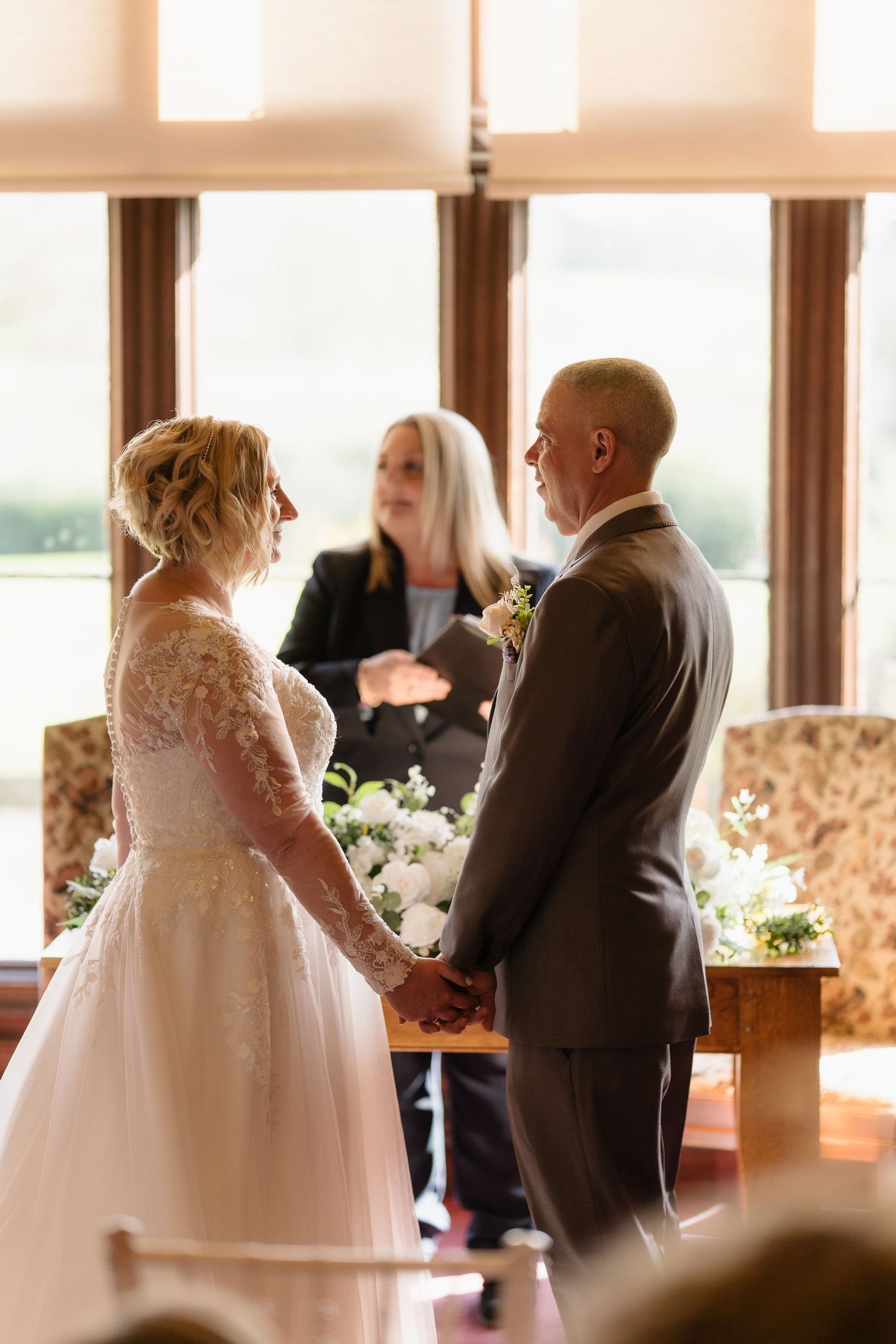 A bride and groom hold hands at the altar during their wedding ceremony, officiated by a woman in a sunlit room.