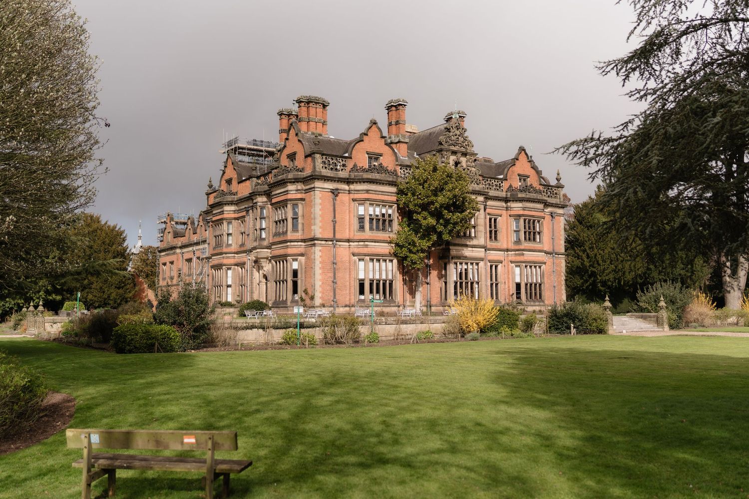 A grand Victorian red-brick mansion with manicured lawns, ornate chimneys, and a wooden bench in the foreground under a cloudy sky.