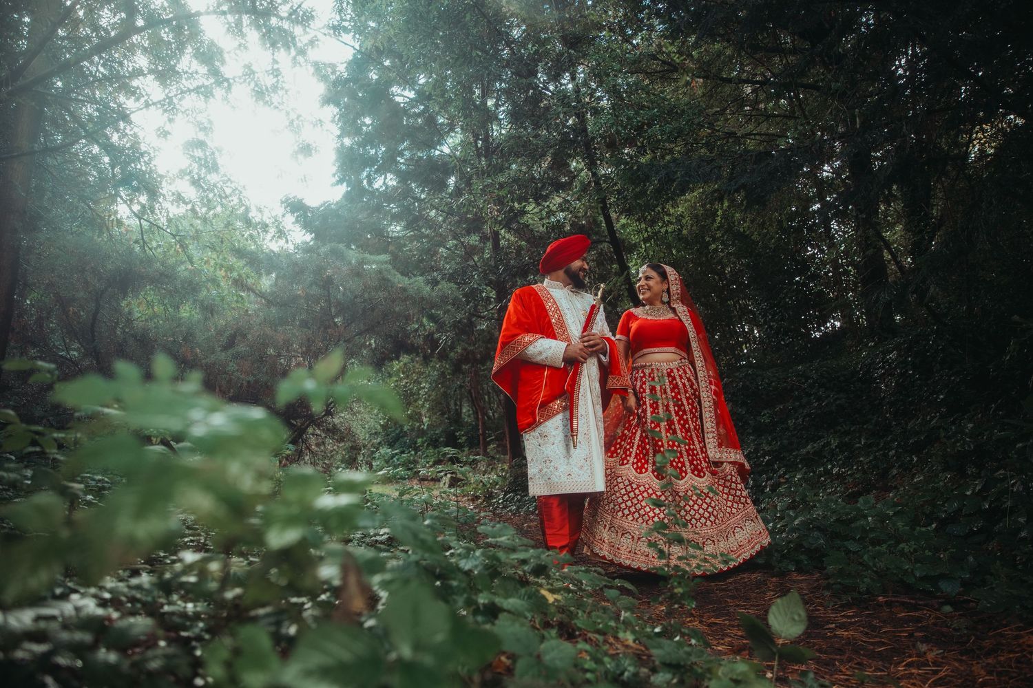 Indian bride and groom in traditional red wedding attire walking through a forest path.
