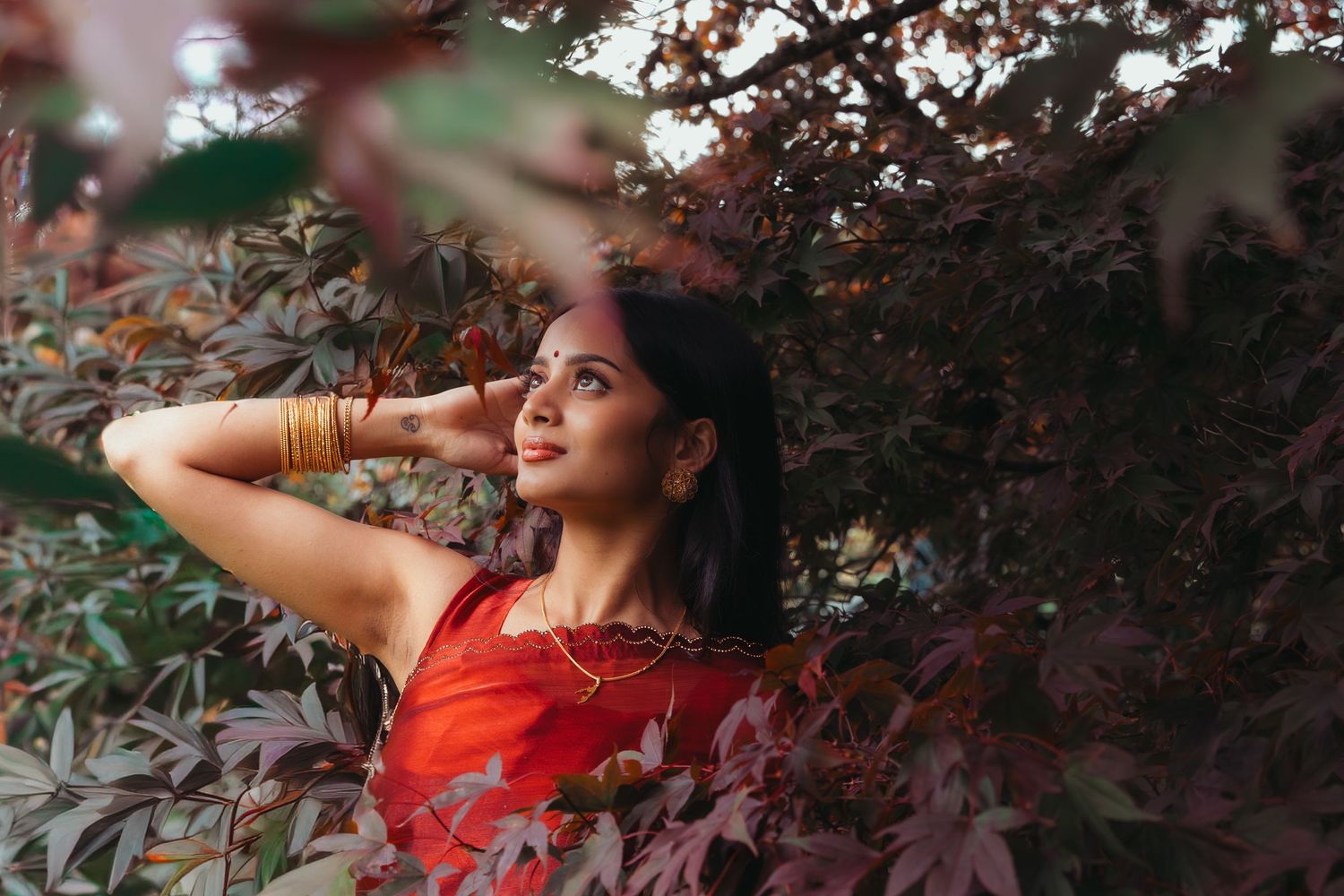 A smiling woman in a red saree poses among autumn red-leafed trees with gold bangles.