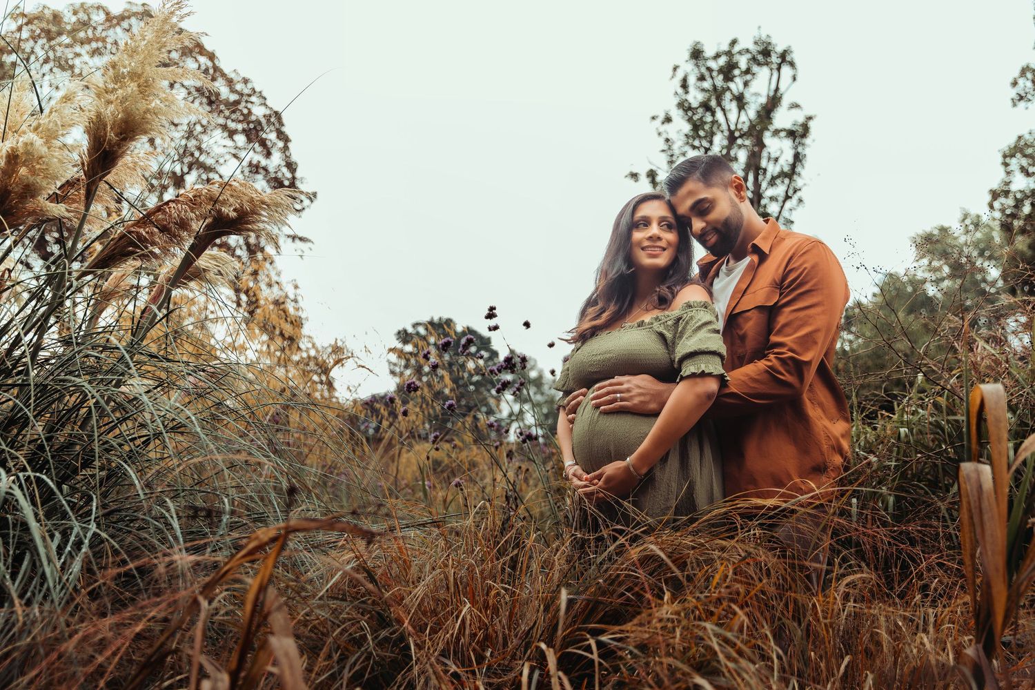 Pregnant couple embracing in an autumn field, man holding baby bump surrounded by tall grass.