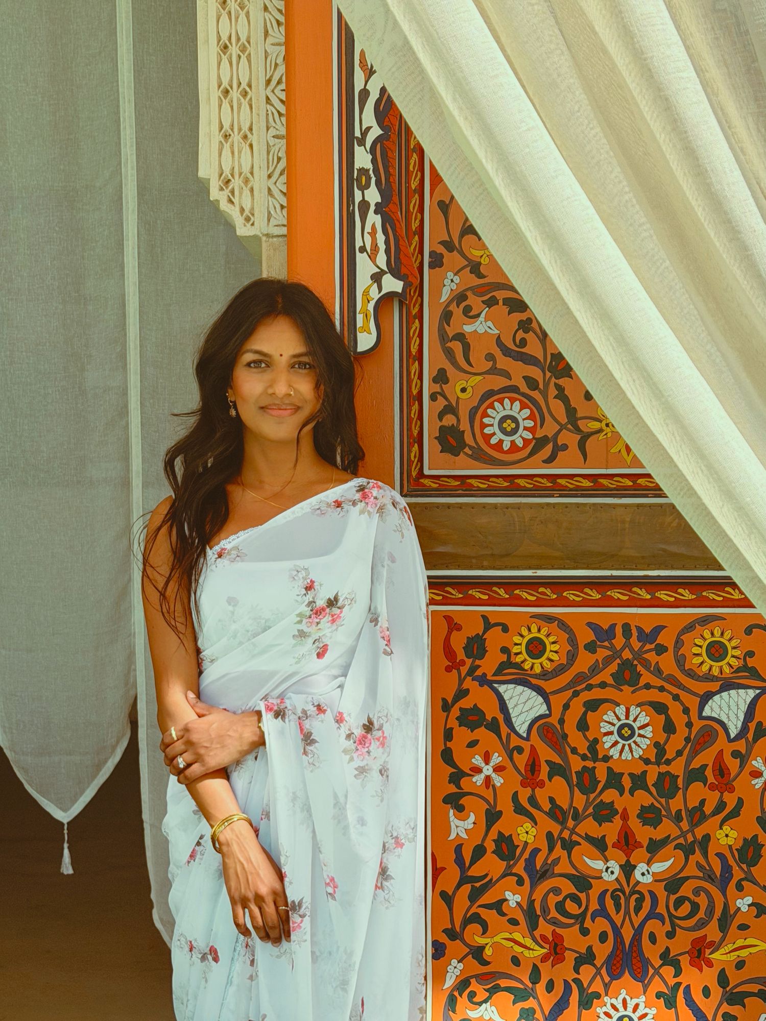 A woman in a light floral saree stands by ornate colorful Indian architectural decor.