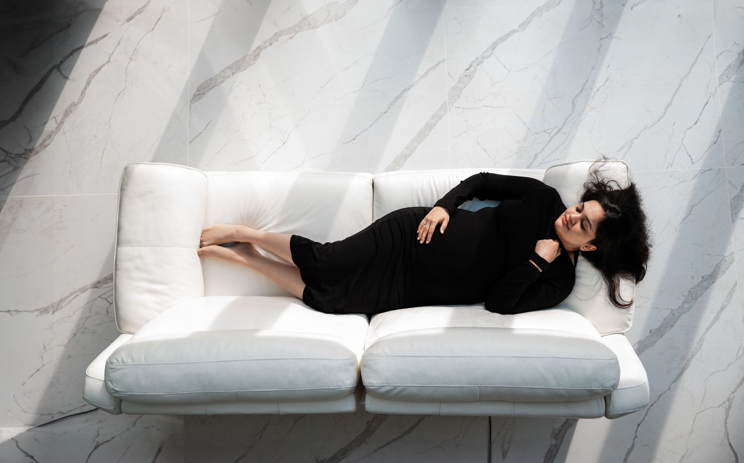 A woman in a black dress reclines gracefully on a white sofa against a marble wall.