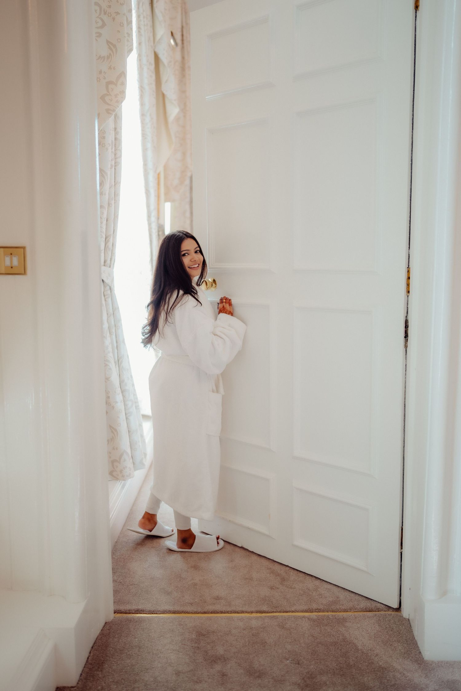 Woman in white bathrobe and slippers standing by a bright window, smiling and holding a drink indoors.