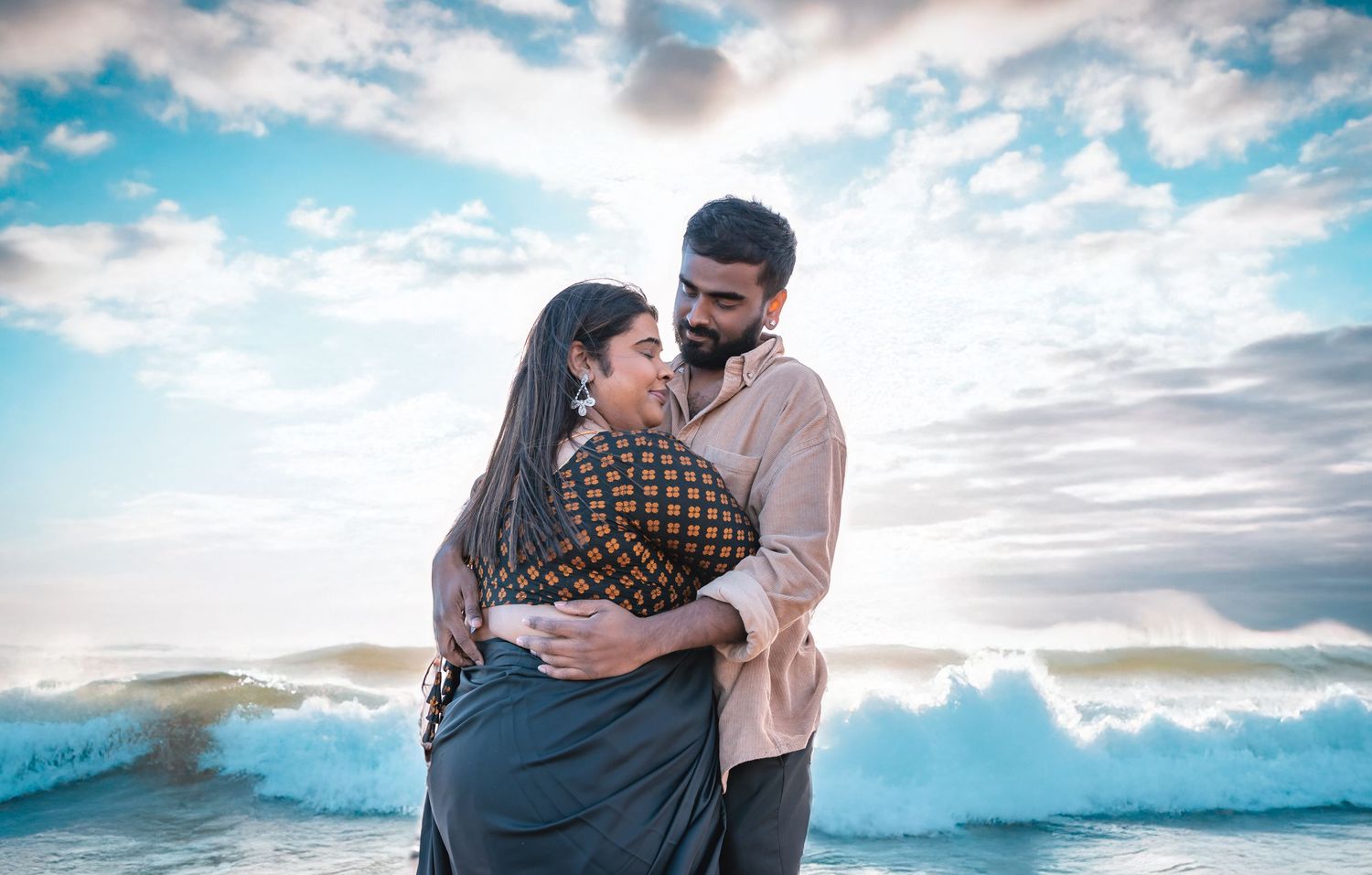 Couple embracing on a beach with crashing waves, woman in saree and man in casual shirt.