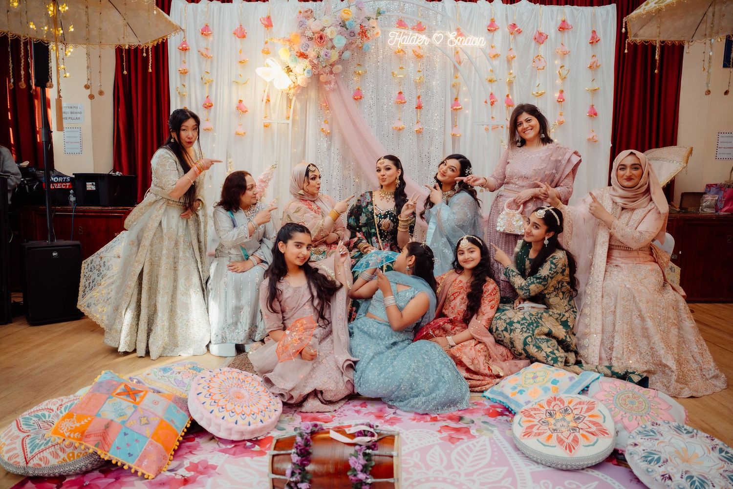 Group of women in colorful South Asian attire celebrating at a festive indoor mehndi ceremony with floral decorations.