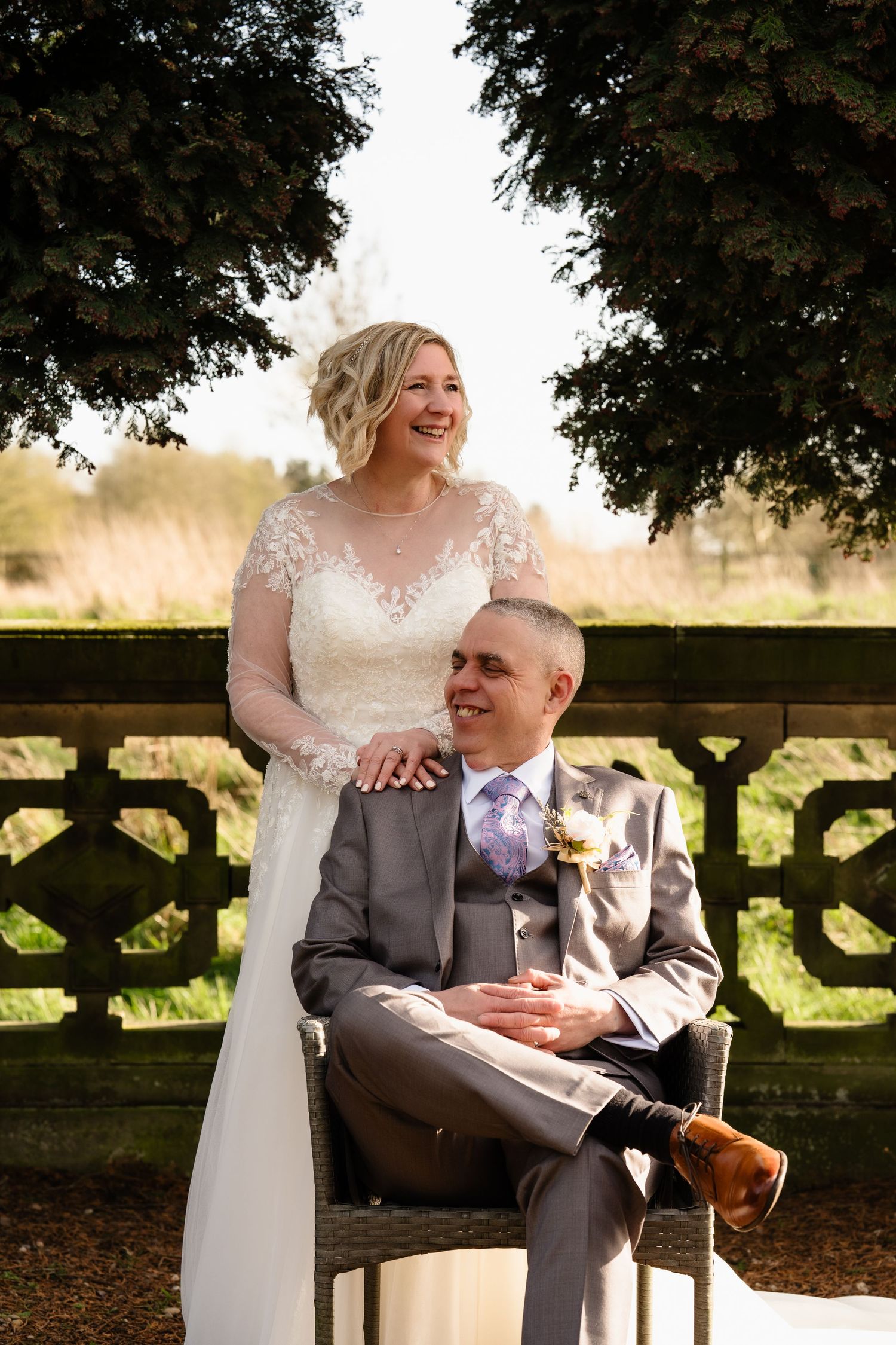Bride in lace gown stands behind seated groom in gray suit, both smiling, in an outdoor garden setting.