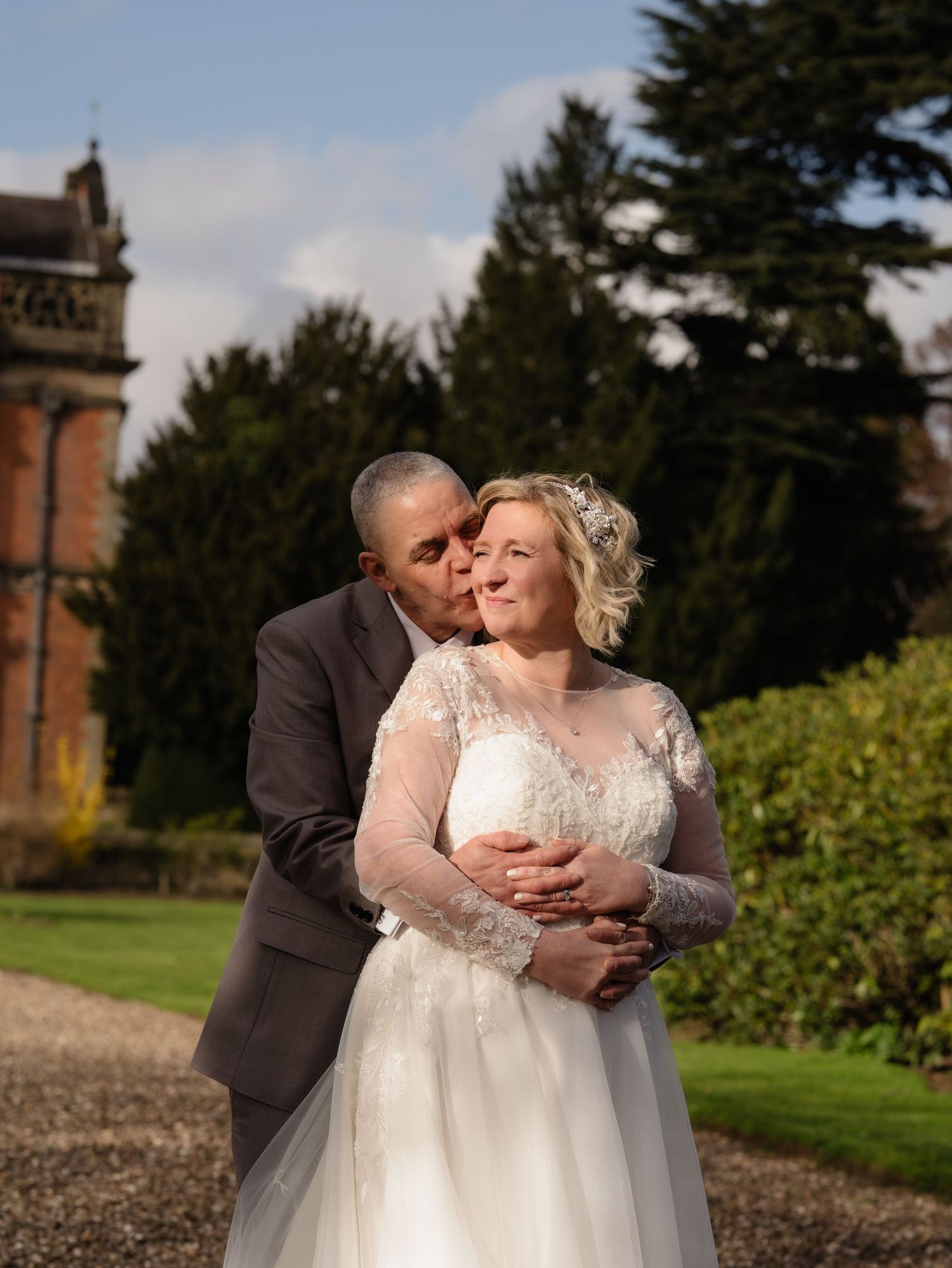 A groom in a gray suit kisses his bride in a lace wedding gown outdoors near a grand estate.