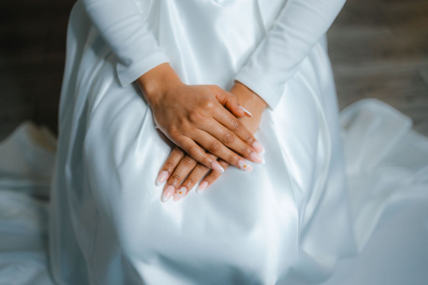 Close-up of bride's hands with elegant manicure resting on white wedding dress.