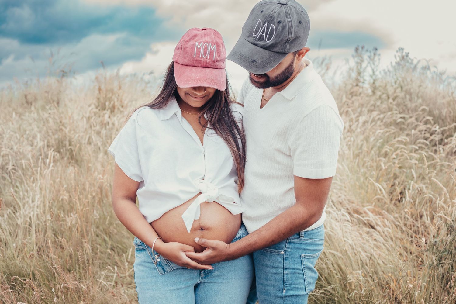 Expecting couple in matching white outfits and caps cradling baby bump in a golden field.