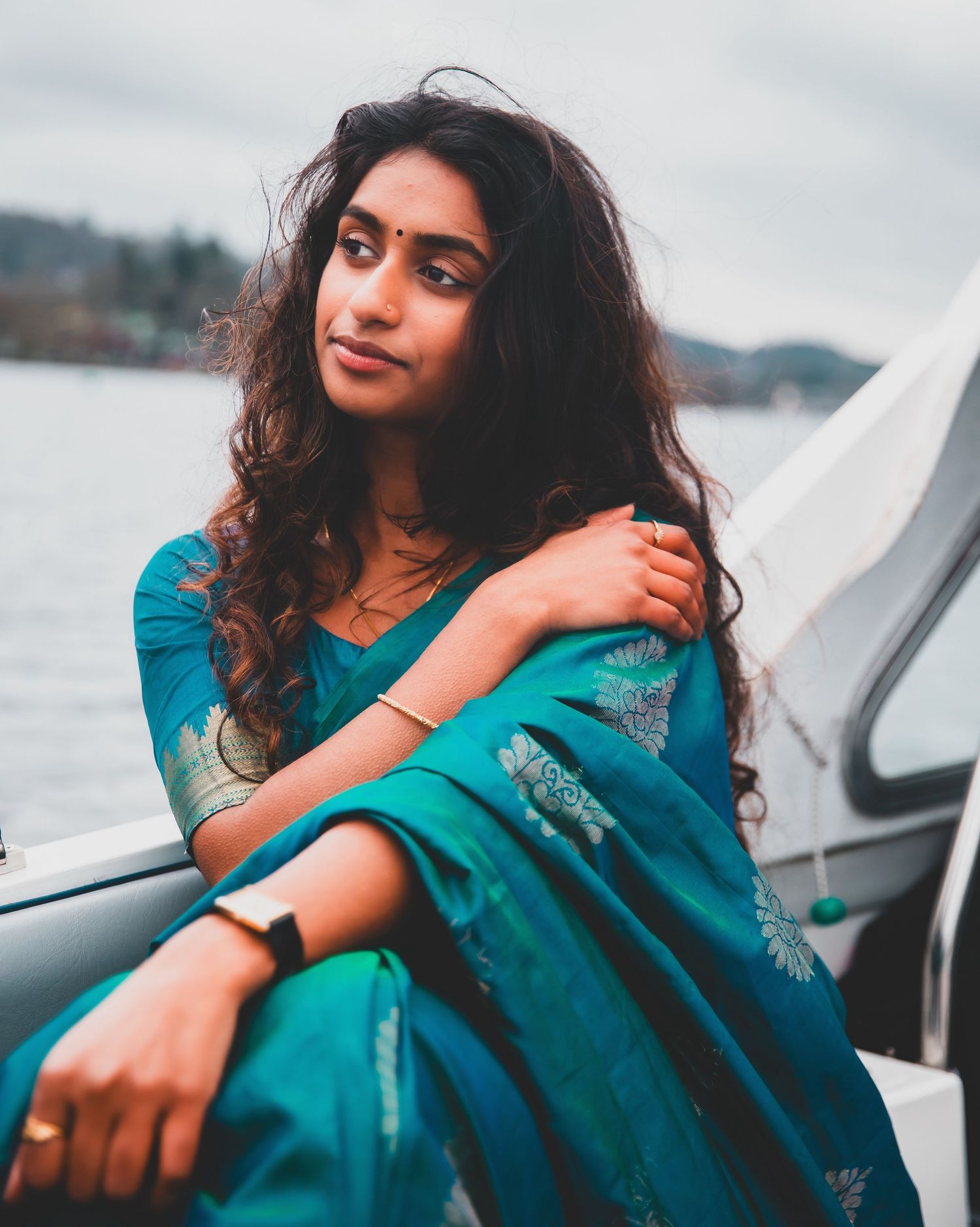 A young woman in a teal embroidered saree sits on a boat with a lake backdrop.