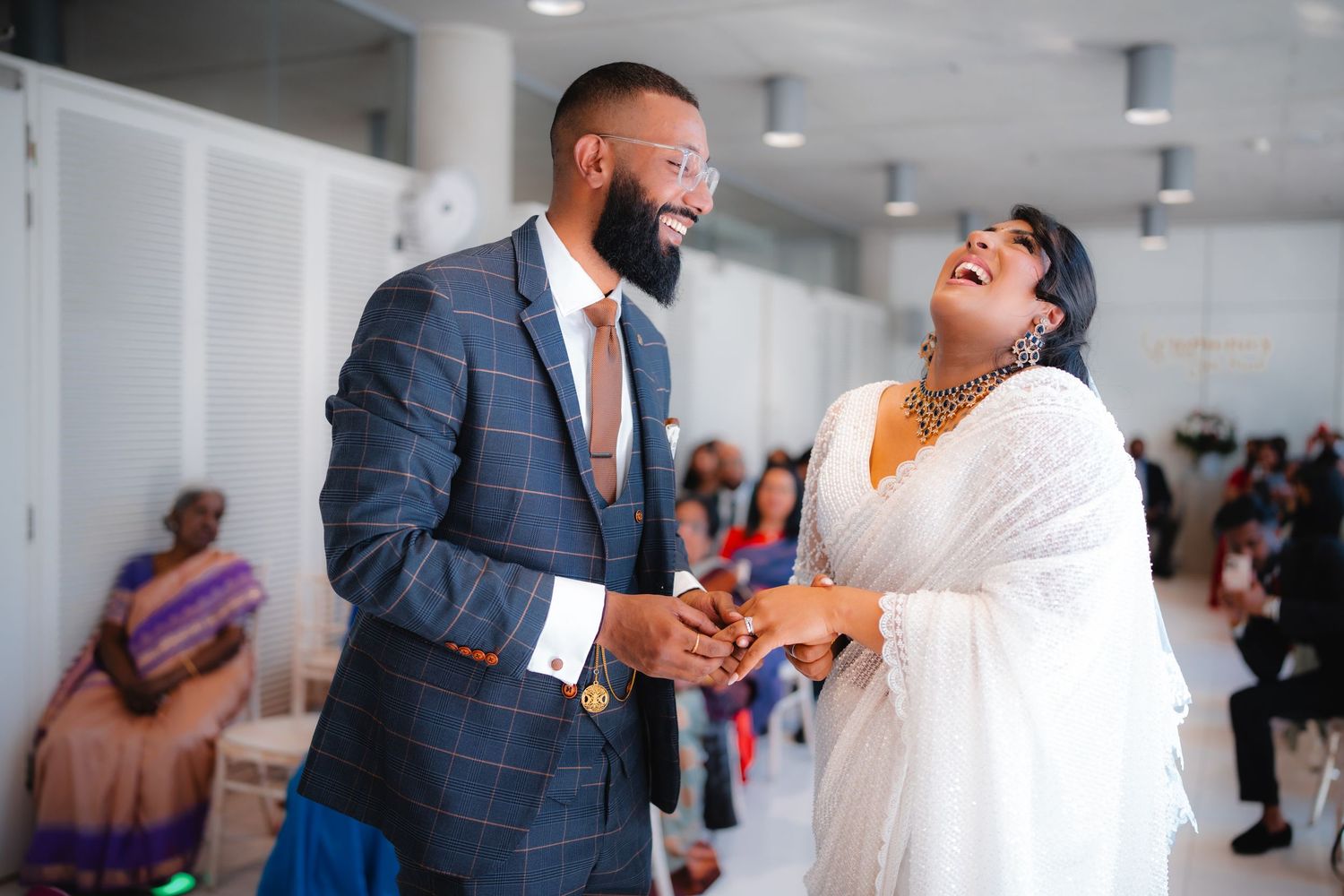 A joyful couple in elegant attire laughing together at a wedding celebration, man in plaid suit and woman in white dress.