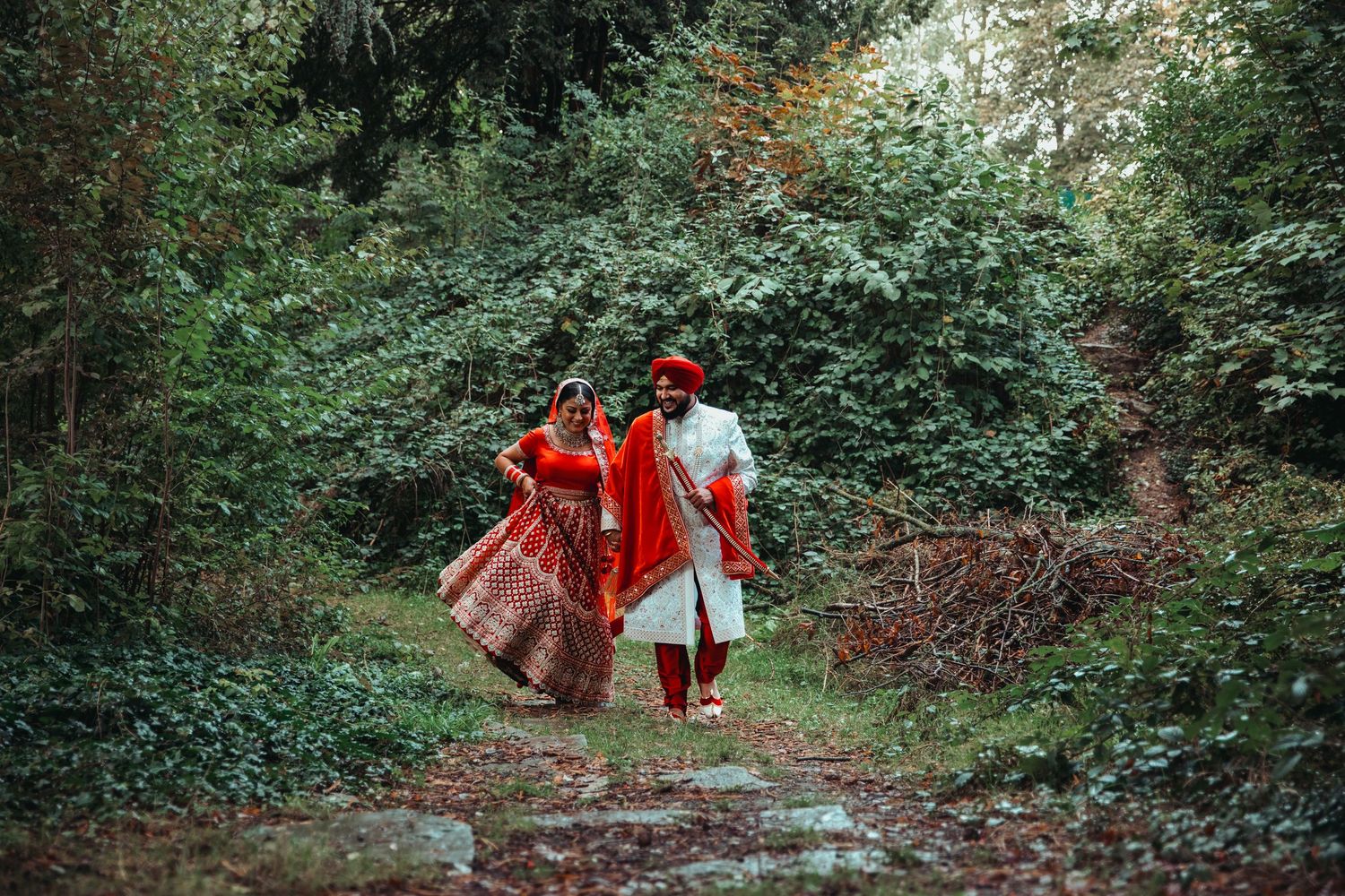 Indian couple in traditional red wedding attire walking hand in hand along a lush green forest path.