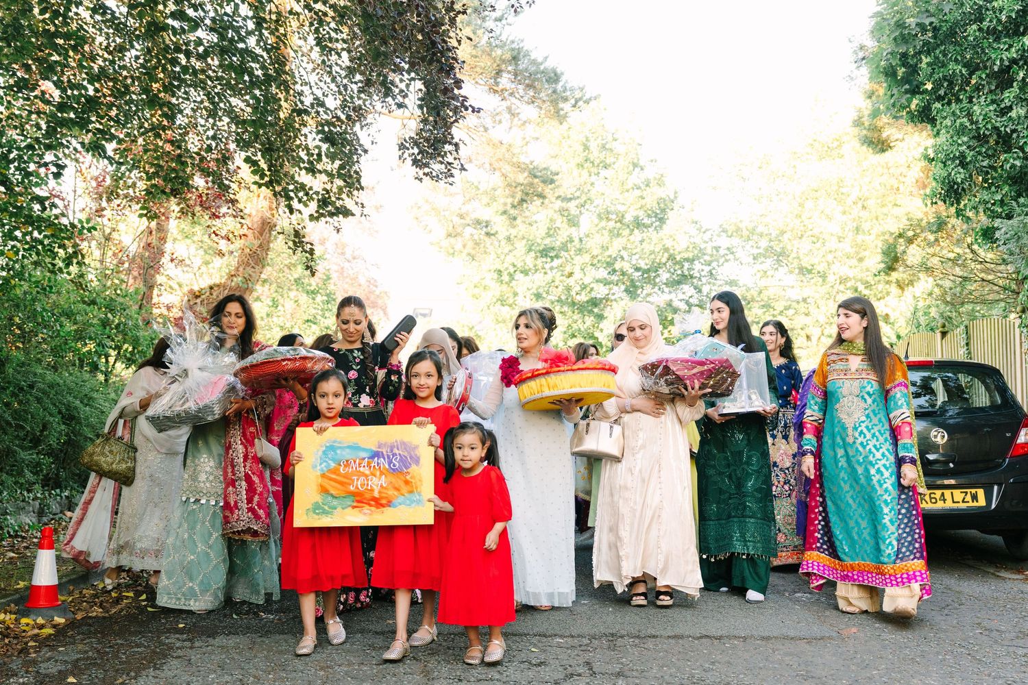 A group of women and girls in colorful traditional attire pose outdoors, holding gifts and flowers at a festive cultural celebration.
