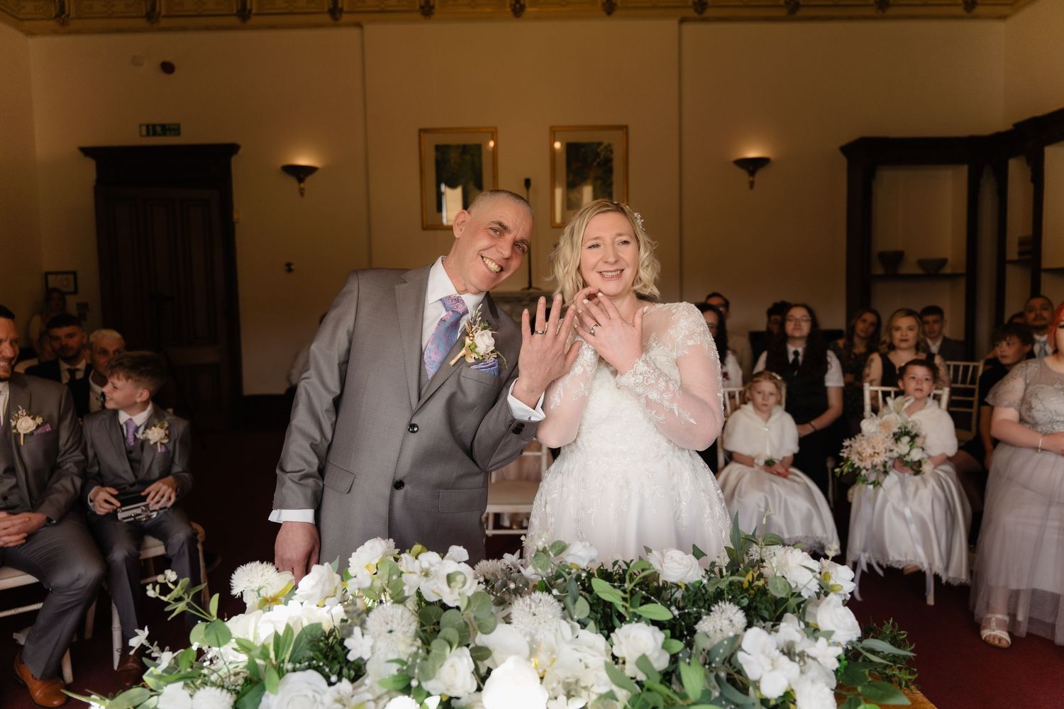Newlywed couple smiling and showing wedding rings at ceremony table with white floral arrangements and guests seated behind.