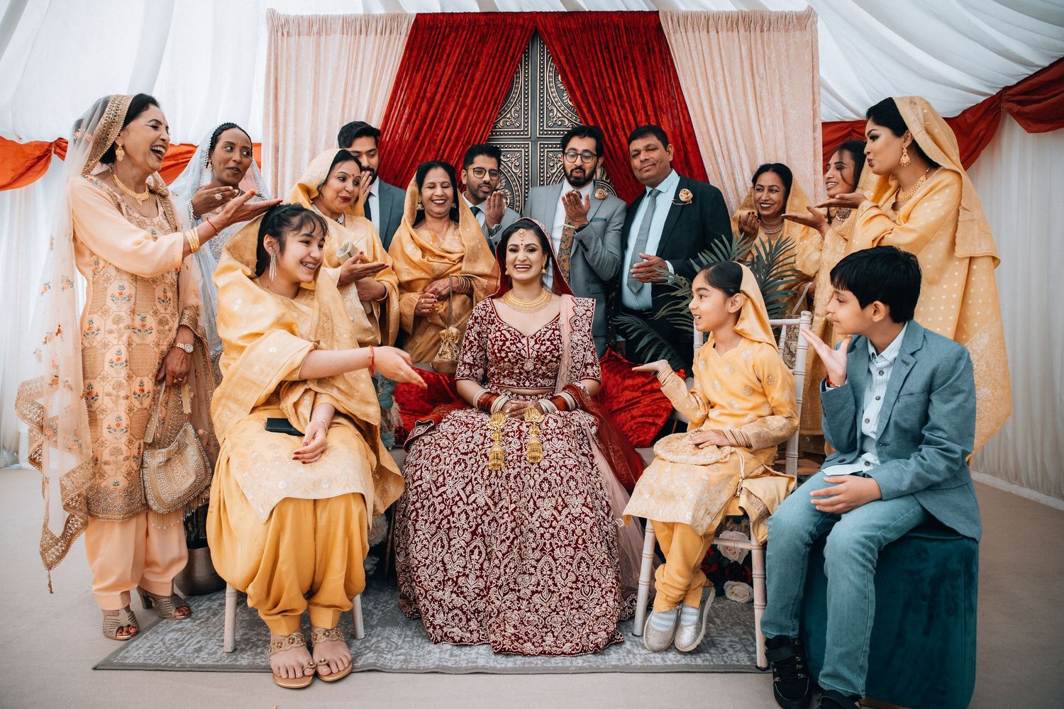 Indian bride in red lehenga surrounded by smiling family members in matching golden outfits.