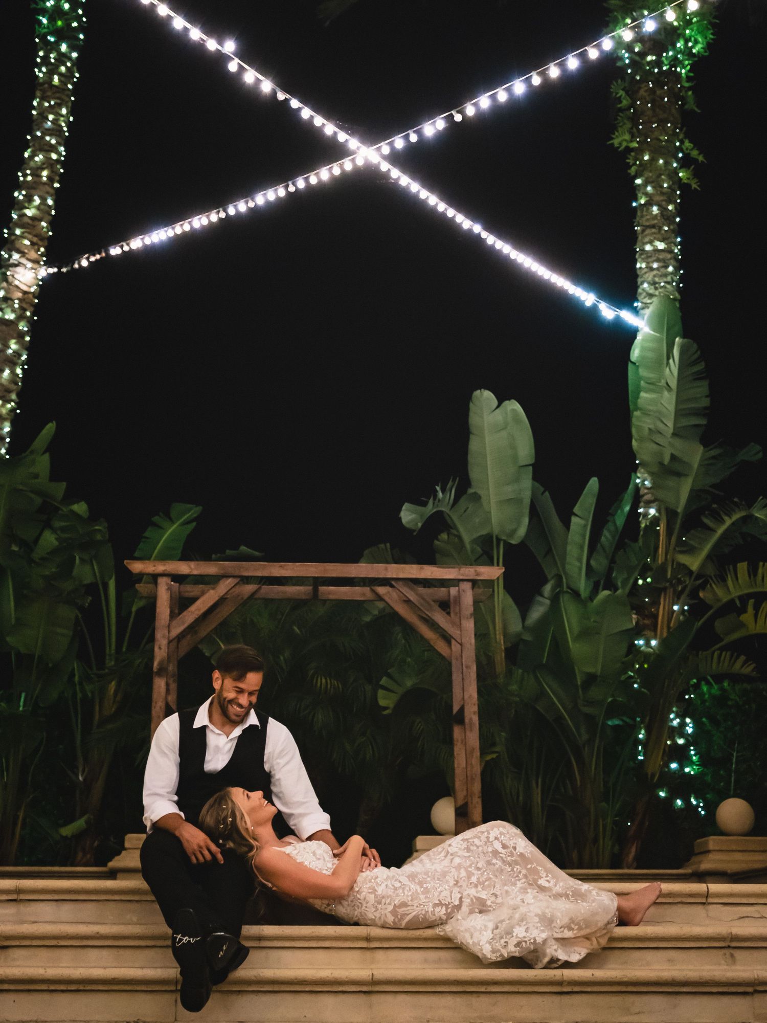Bride and groom relax under fairy lights at an outdoor nighttime wedding reception.